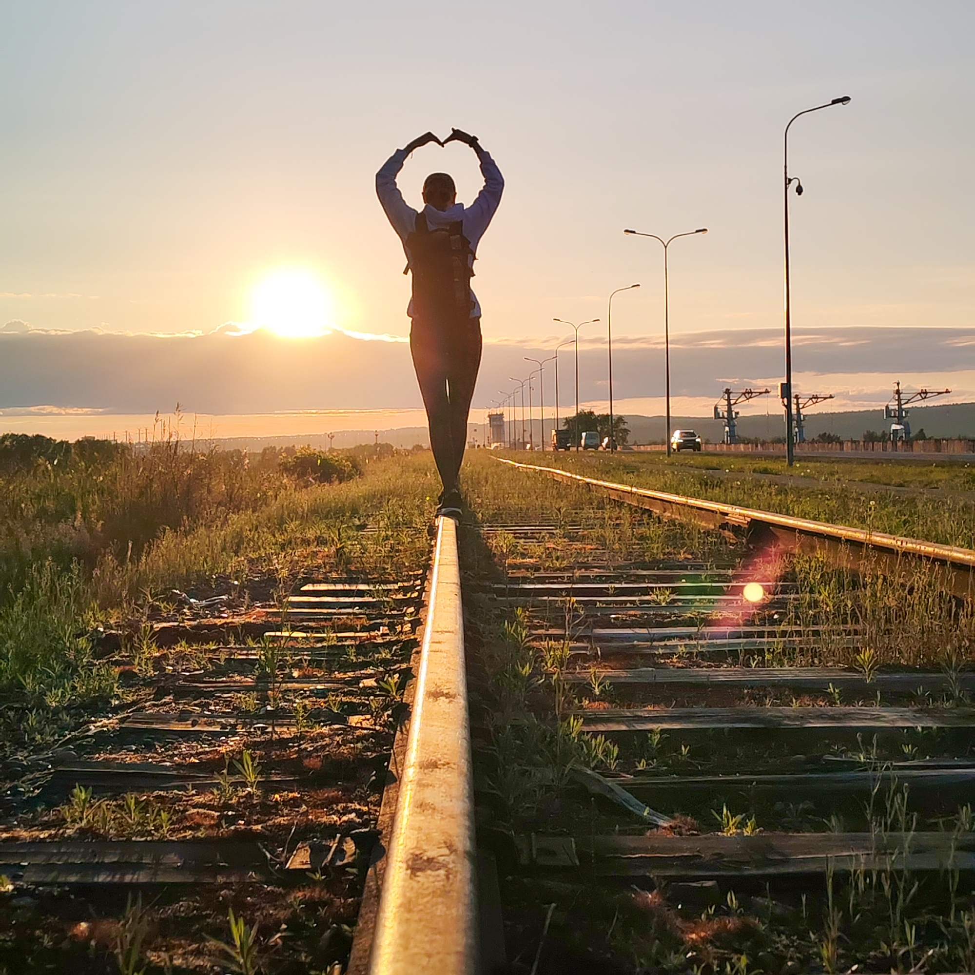 The image captures a serene sunset scene along a set of railroad tracks in Chaikovsky, Russia. A lone figure, viewed from behind and silhouetted against the bright sun, stands precariously balanced on one of the metal rails. The person, wearing a light-colored top and dark pants with a backpack, has their arms raised above their head, hands joined to form a heart shape, conveying a sense of love, peace, or appreciation for the moment.
The sun, a brilliant orb, is low on the horizon, casting a warm, golden glow across the sky and illuminating the edges of scattered clouds. The sky transitions from a bright yellow and orange near the sun to a soft, pale blue higher up. The railroad tracks, partially overgrown with vibrant green grass and weeds, stretch into the distance, appearing disused or less frequently traveled. Wooden sleepers are visible beneath the tracks.
In the mid-ground and background to the right, several tall streetlights or utility poles stand in a line, creating further silhouettes. Beyond them, some distant, possibly industrial, structures or gantry cranes can be vaguely discerned, along with what might be cars on a parallel road. The overall impression is one of quiet contemplation and natural beauty meeting human infrastructure at the end of the day. There is no visible text in the image.