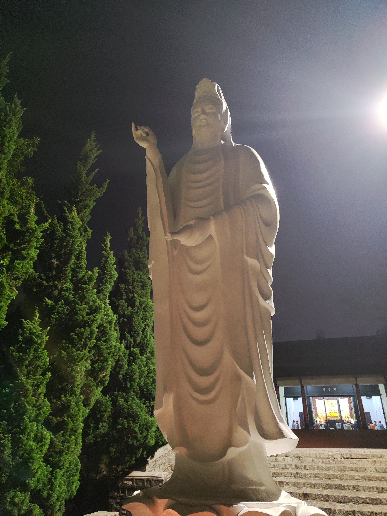 A nighttime, upward-angle shot depicts a tall, white statue of a Buddhist figure positioned on a tiered platform, likely Quan Yin, adjacent to tall, green, cypress-like trees on the left. The statue has its right hand raised with fingers extended and its left hand holding an object. To the right, behind the statue, a building with visible interior lights and people inside sits atop a set of stone steps. The scene is illuminated by a light source above and to the right of the statue. The location is speculated to be the Linh Phuoc Pagoda in Da Lat, Vietnam.