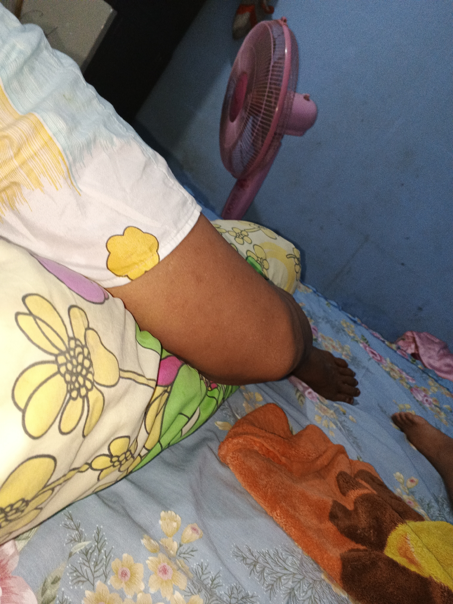 The image shows a close-up view of a person's arm and legs lying on a bed. The bed is covered with a blue floral-patterned sheet and has several pillows. One pillow has a large yellow flower design, while another has a white background with yellow and green floral patterns. An orange towel with a brown and yellow design is draped over the bed. A pink electric fan is visible in the background, mounted on a blue wall. The scene appears to be indoors, likely a bedroom. The lighting suggests it might be daytime or under artificial light.
