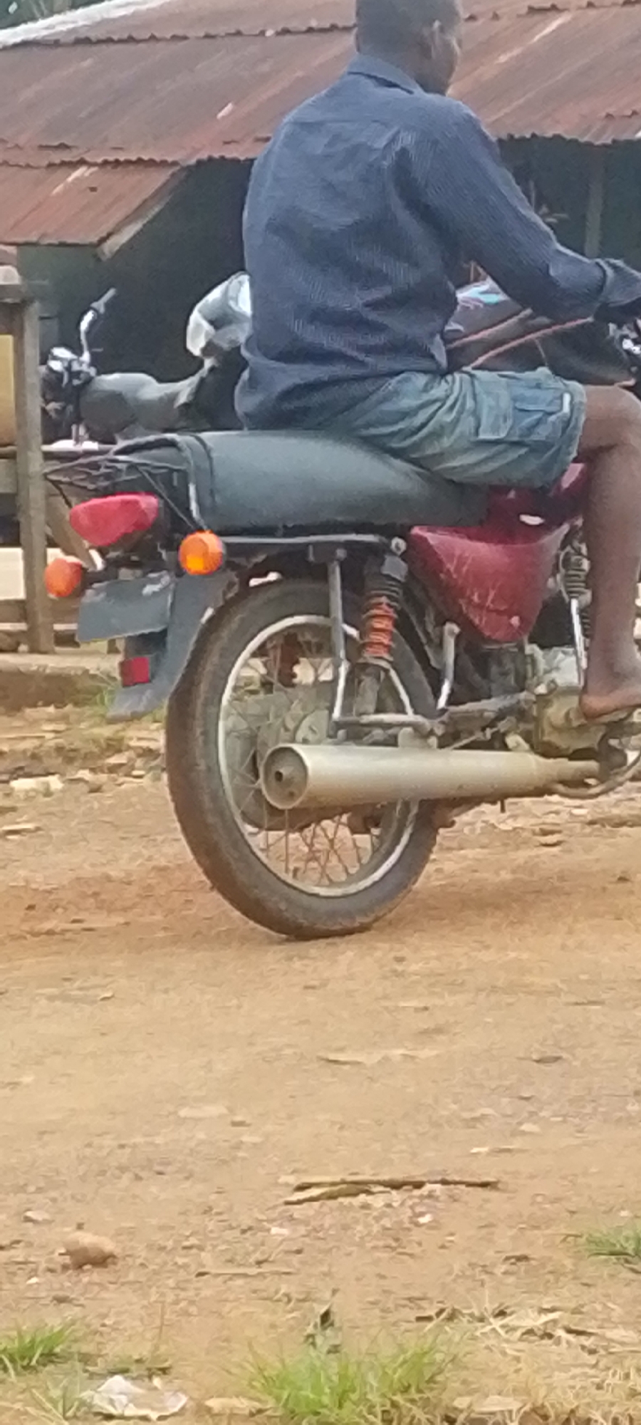 The image shows a person sitting on a red motorcycle, facing away from the camera. The person is wearing a dark blue striped shirt and light blue shorts. Their right leg is visible, with a bare foot. The motorcycle has a black seat, a black rack on the back, and a red tail light with two orange turn signals. The rear wheel, spokes, and exhaust pipe are clearly visible. The motorcycle is parked on a dirt road. In the background, there is a building with a corrugated metal roof and a wooden structure. The scene appears to be outdoors, possibly in a rural or semi-urban area. The lighting suggests it could be daytime, with diffused light, possibly overcast or shaded. There are no visible signs or text in the image. The overall impression is that of a person preparing to travel or taking a break on their motorcycle in a casual setting.