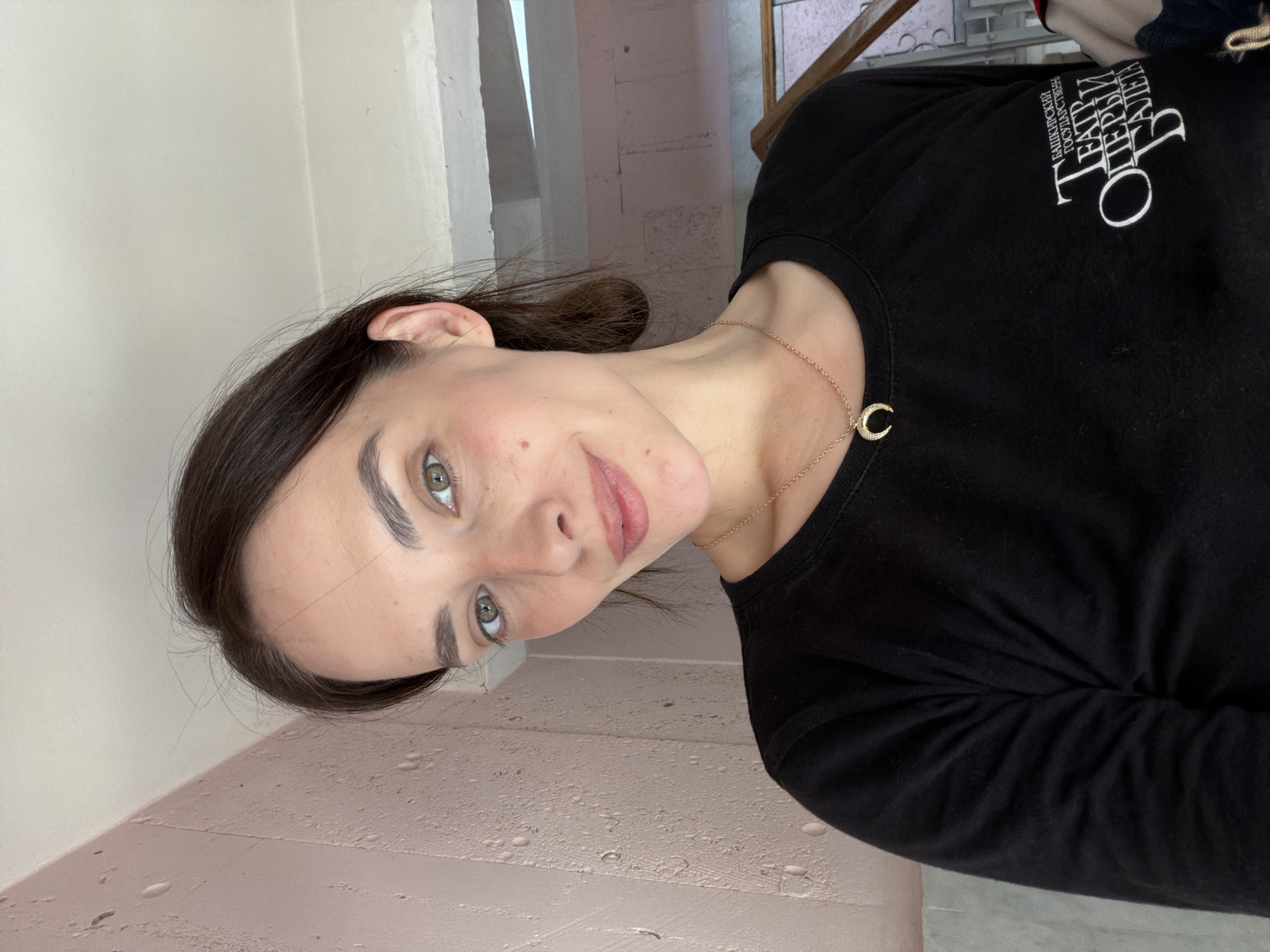 A young woman with dark hair and green eyes smiles at the camera, her head tilted to the side. She is wearing a black long-sleeved shirt with a gold necklace featuring a crescent moon pendant. The shirt has white text on the upper left side. The text appears to be in Cyrillic script and reads "БАШКИРСКИЙ ГОСУДАРСТВЕННЫЙ ТЕАТР ОПЕРЫ И БАЛЕТА". The background suggests an indoor setting, possibly a hallway or backstage area, with pale pink walls and architectural details visible. The lighting appears to be artificial, casting soft shadows. The image is oriented with the top of the woman's head towards the right side of the frame.