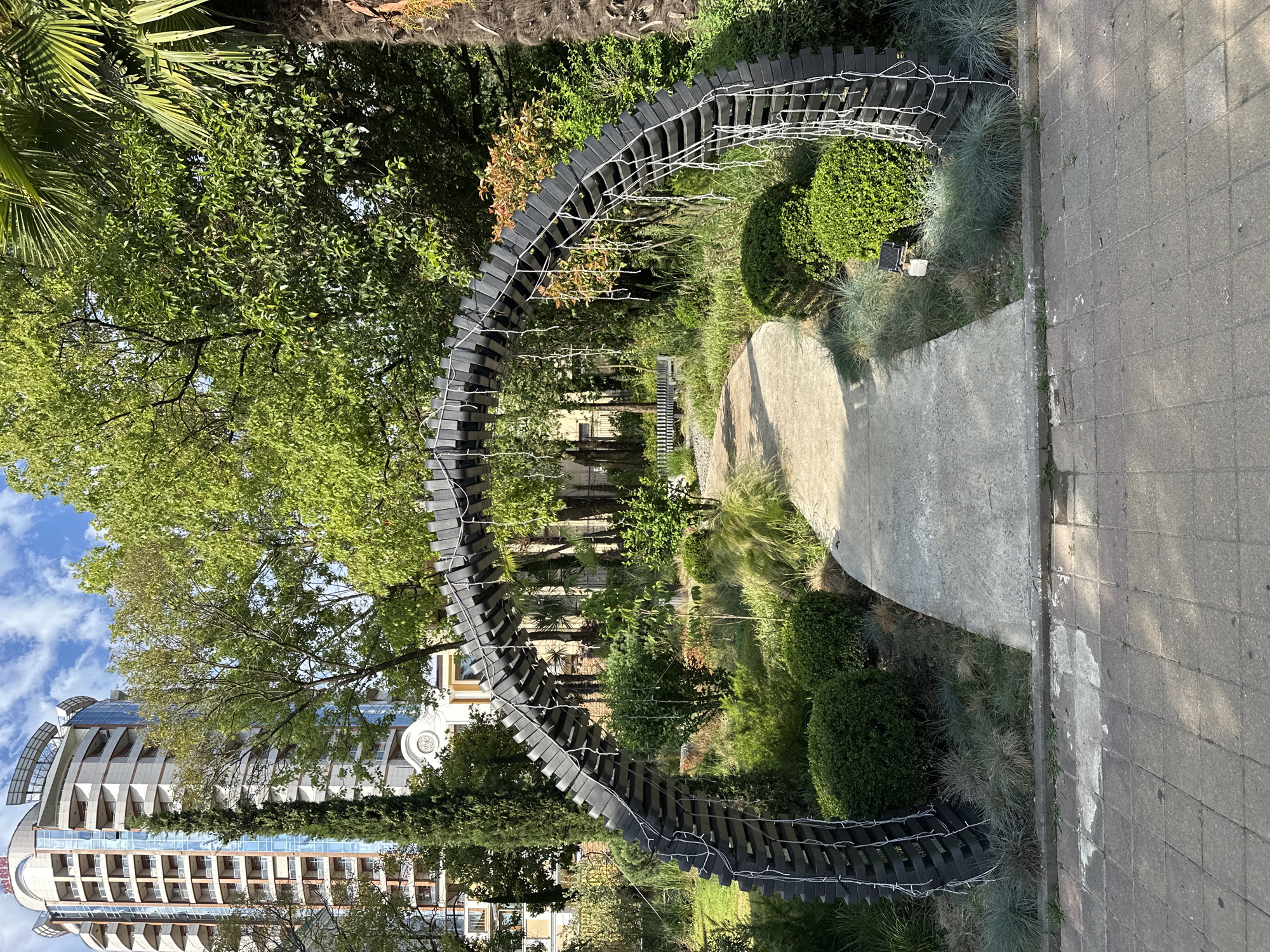 The image depicts an outdoor scene in Sochi, Russia, likely within a park or garden area. A prominent arched structure, constructed from dark, interlocking segments and adorned with white string lights, spans a paved pathway. The pathway leads into a verdant landscape featuring manicured bushes, ornamental grasses, and various trees, including a tall cypress to the left and a palm tree to the right. In the background, a modern, multi-story building with a curved facade stands tall, its windows reflecting the sky. The sky is a bright blue with scattered white clouds. There are no people visible in the image. The overall environment suggests a well-maintained public space on a clear day.