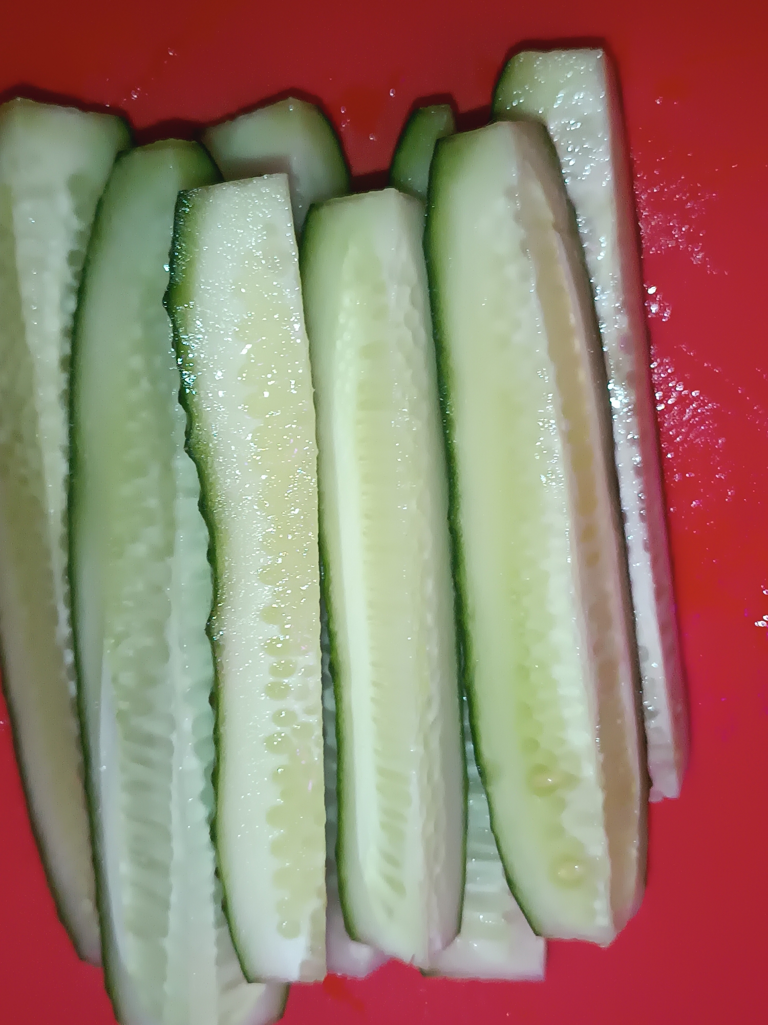 The image displays several sliced cucumbers arranged vertically on a red surface. The cucumbers are cut lengthwise, revealing their pale green flesh and seeds. The cut edges appear moist and slightly textured. The red background is solid and appears to be a flat surface, possibly a cutting board or a plate. There is no indication of a specific location in the city of Sluck, Belarus, or any people, actions, or interactions depicted. The image focuses solely on the prepared cucumbers.