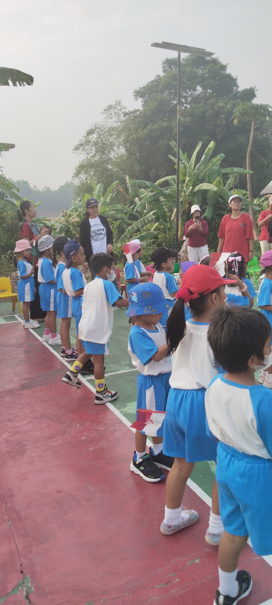 In Taban, Indonesia, a group of children, dressed in blue and white uniforms, stand in a line on a red and green court, likely a sports field or playground. Many of the children are wearing hats, some with colorful designs, and others are holding small flags. Adults are present, observing the children. In the background, lush green trees, including banana plants, are visible, and a tall pole with a solar panel stands against a hazy sky. The scene suggests an outdoor activity or event involving young children.