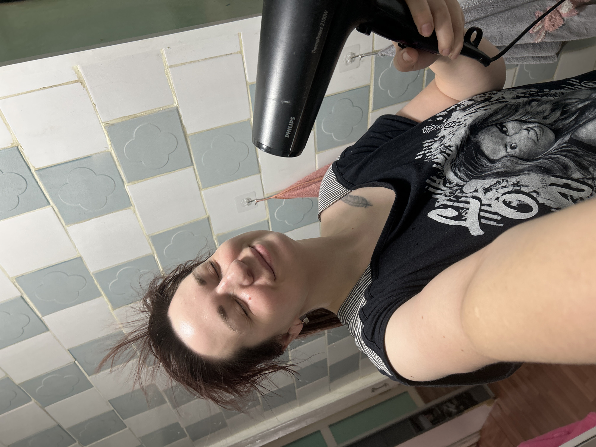 A woman is drying her hair with a Philips hairdryer. She is wearing a black t-shirt with a silver graphic. The location appears to be a bathroom with white and blue tiled walls. The city of Фоки, Russia cannot be determined from the image.