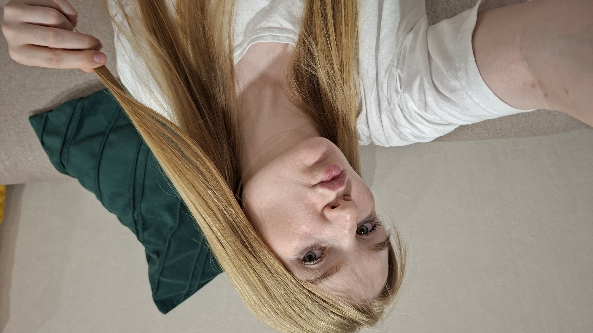 A young Caucasian woman with long, blonde hair and fair skin is seated on a sofa, taking a selfie. She is wearing a white t-shirt and has her lips pursed in a kissing gesture. A dark green, textured pillow is visible behind her. The background appears to be a wall, with a hint of yellow on the right side of the frame. The lighting suggests an indoor setting. There are no discernible landmarks or contextual clues to pinpoint a specific location within Chaikovsky, Russia.
