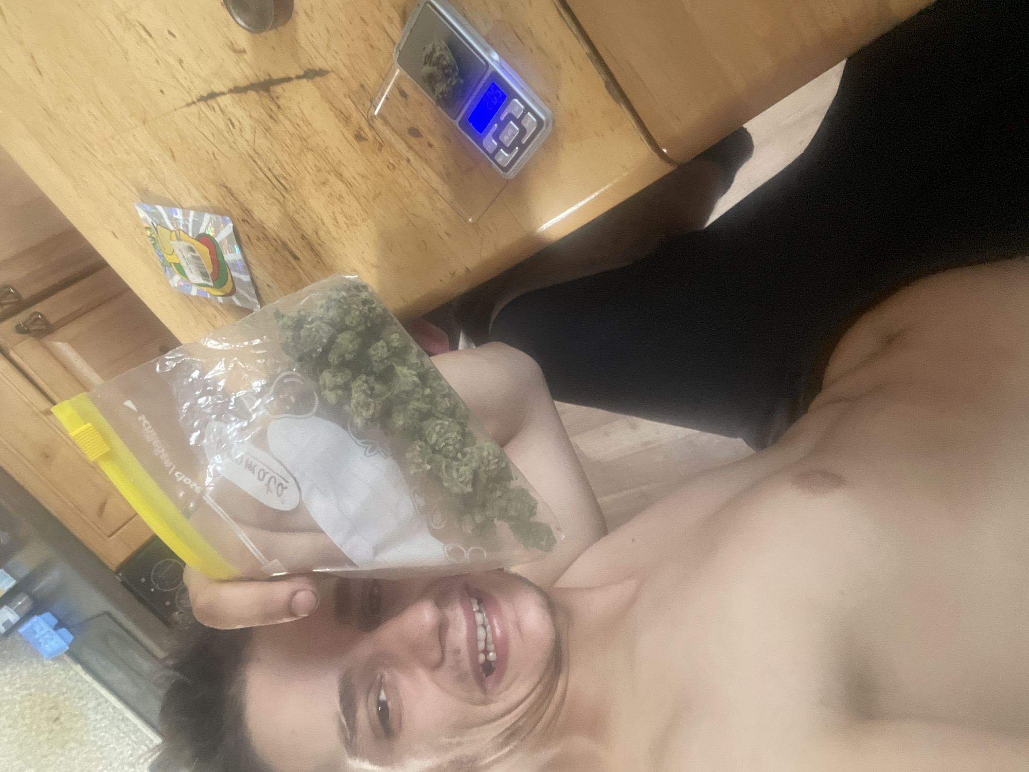 The image shows a person lying on the floor, holding a bag of marijuana. On a wooden table in the background, there is a digital scale with a bud of marijuana on it, displaying "2.05". There is also a sticker with a colorful, cartoonish character. The person is smiling and appears to be in good spirits. The lighting suggests it may be daytime, but it is difficult to determine the exact time. The overall scene appears to be indoors, likely in a casual setting.
