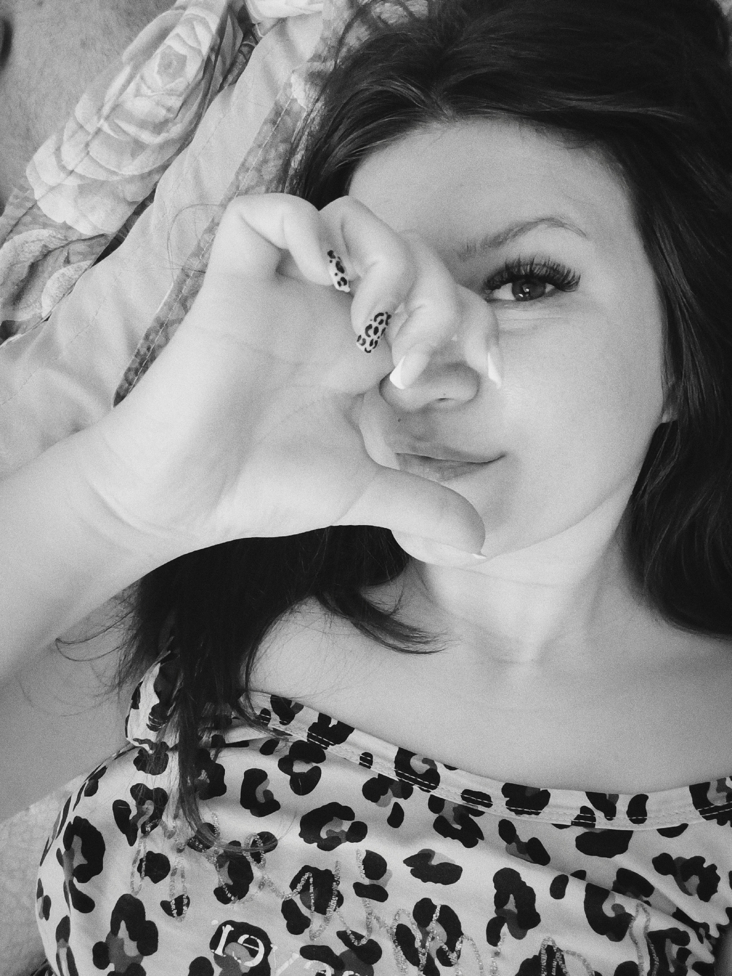 A woman is lying down, presumably indoors. Her head is tilted to the right, and she is looking directly at the camera. Her right hand is raised, forming a circle with her thumb and index finger. Her ring finger and pinky are also raised. Her nails are painted with a cheetah print pattern. She is wearing a leopard-print tank top, lying on patterned sheets. There is no discernible information to locate this photo in Tobolsk, Russia.