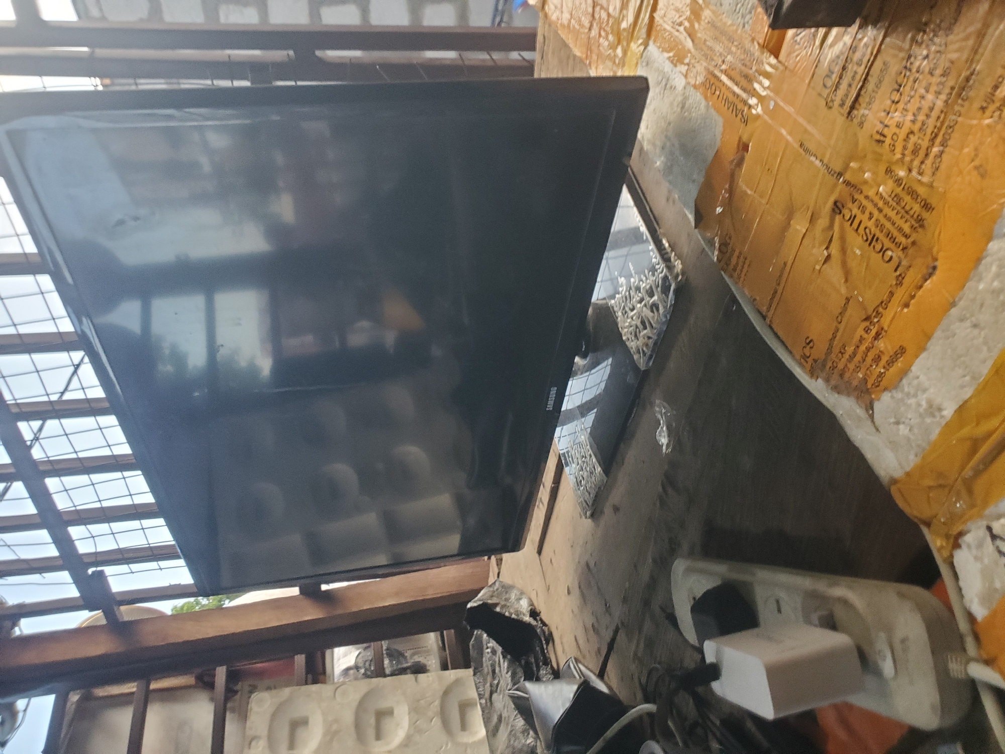 The image shows a Samsung television positioned on a surface next to a cardboard box labeled with logistics information including the city "Guangzhou, China" and text reading "ISAIAH LOGS". An electrical outlet strip with plugs is also visible. The image appears to have been taken outdoors in a cluttered environment, potentially a storage or workshop area. Construction materials such as metal framing and concrete forms are visible. The OCR data suggests the location of a related transaction in B935, Gungzhou, China.