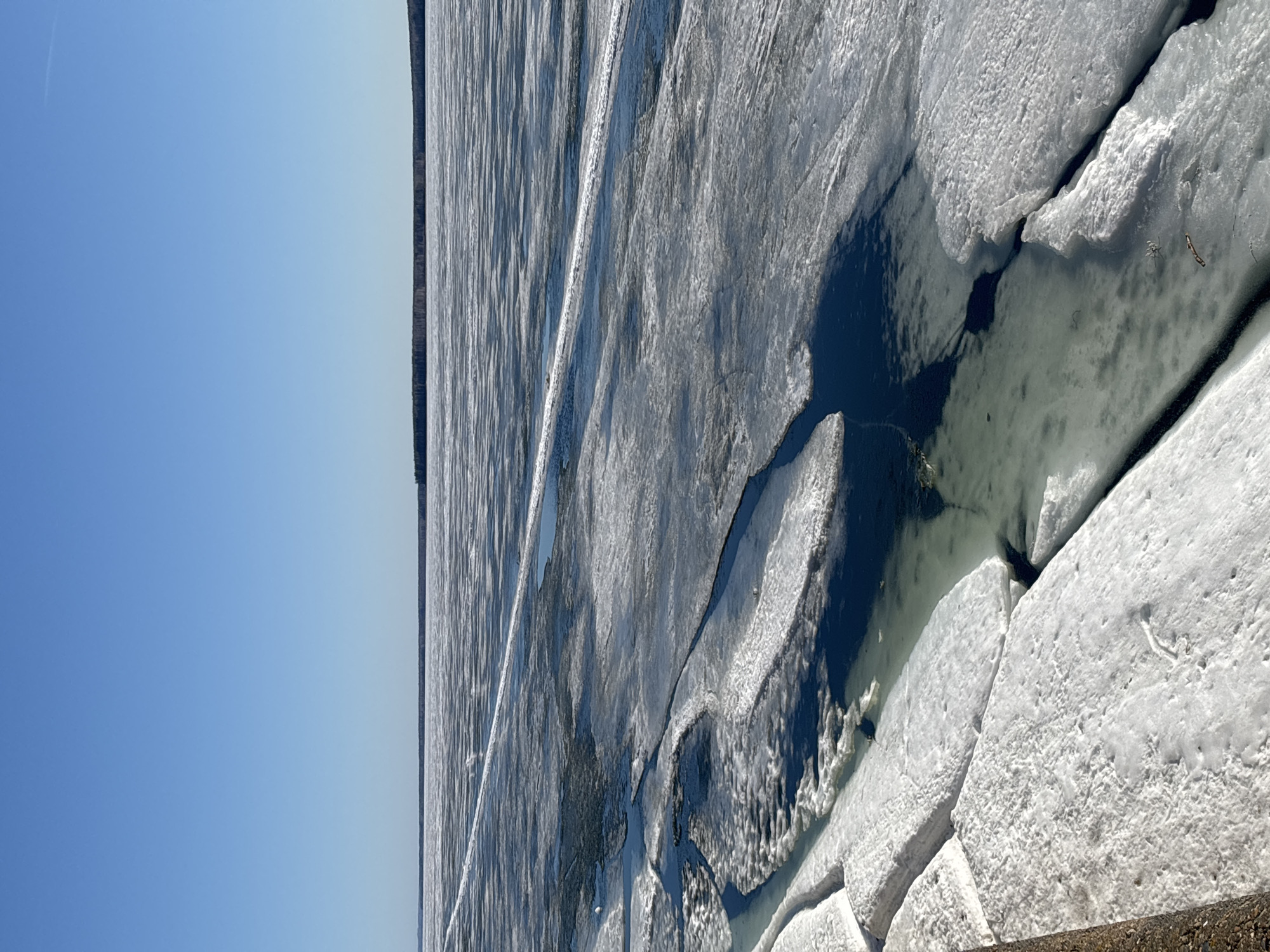 The image depicts a body of water covered in broken ice sheets under a clear blue sky. The edge of the ice is near a tree-lined shore. Based on the location, this could be the Kama River in Chaikovsky, Russia. There are no people visible. The ice is fractured into various sized pieces with some open water visible in the gaps.
