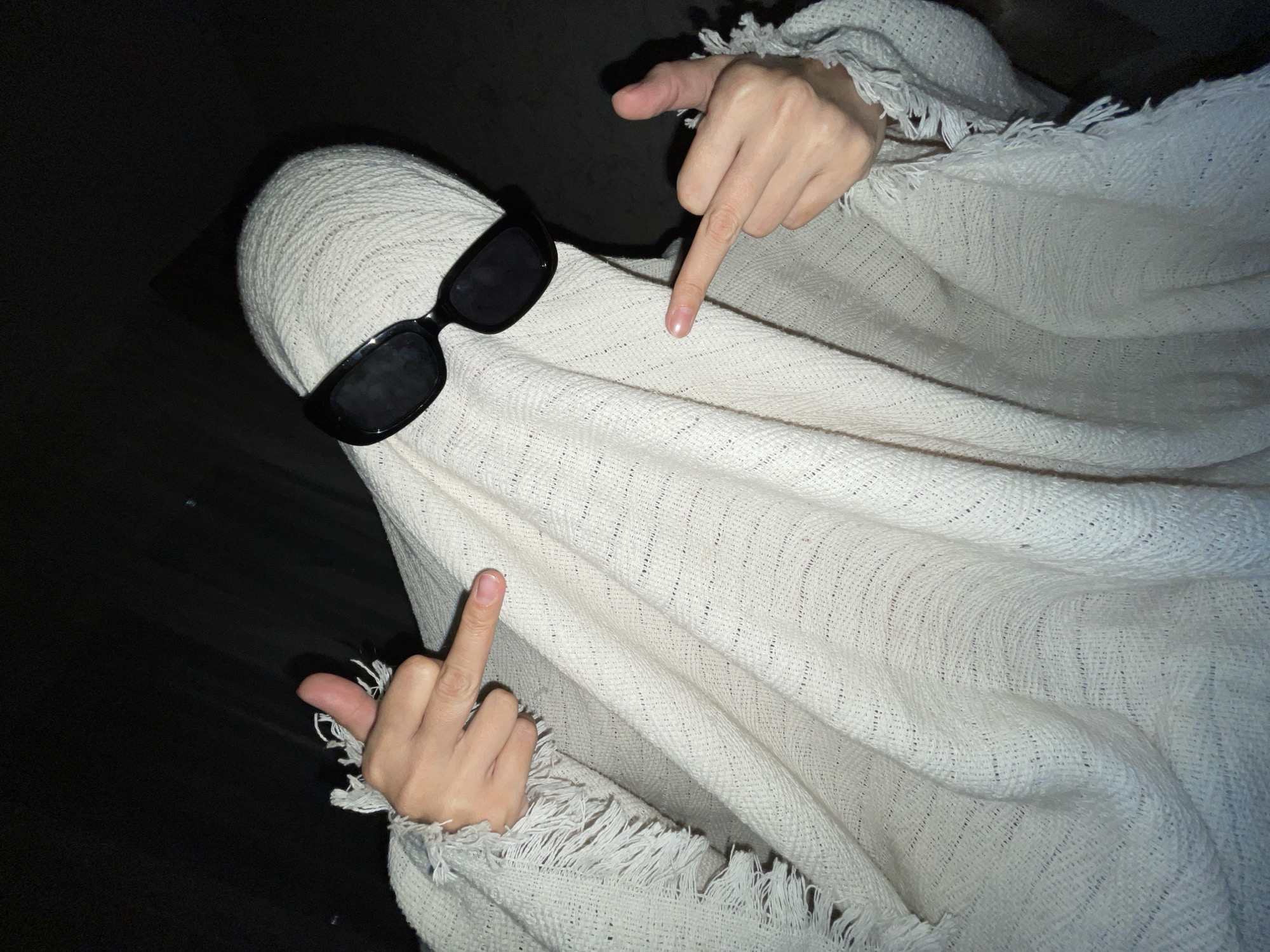 A person dressed as a ghost, wearing a white woven textile as a sheet and black sunglasses, is gesturing with their hands. The left hand is extending the middle finger, while the right hand is pointing downwards. The background appears dark. The location is not apparent from the image.