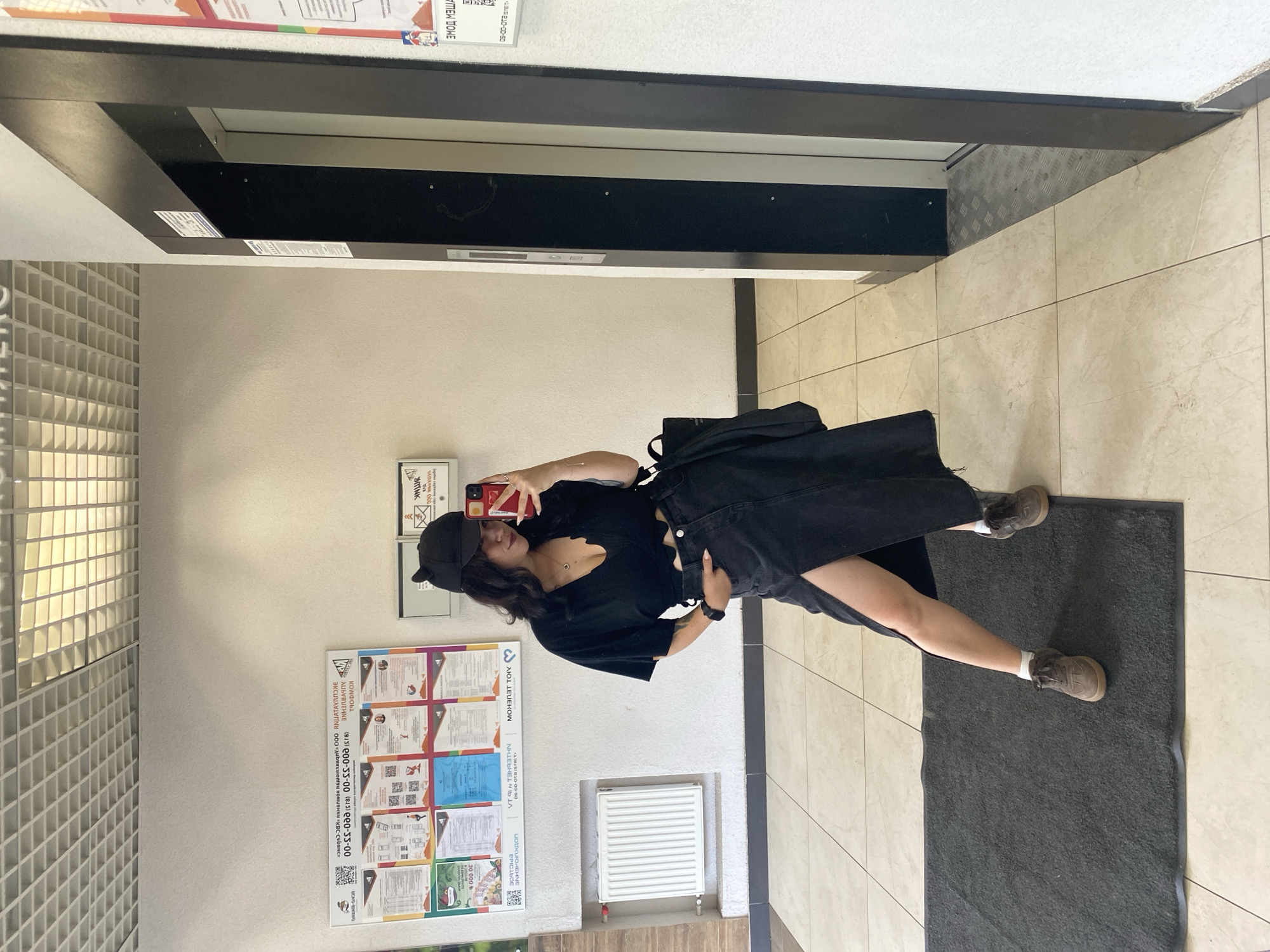 A young woman with dark, wavy hair, wearing a black crop top, a black denim skirt with a high slit, and brown sneakers, stands in a hallway. She is holding a red smartphone and taking a mirror selfie. The hallway has tiled floors, a white wall with bulletin boards displaying various notices and posters, and an elevator to the right. A dark gray mat is on the floor in front of her. The bulletin boards contain text in Russian, indicating the location is likely within a residential building or a communal area in Yanino-1, Russia. The lighting is artificial and appears to be from ceiling fixtures.
