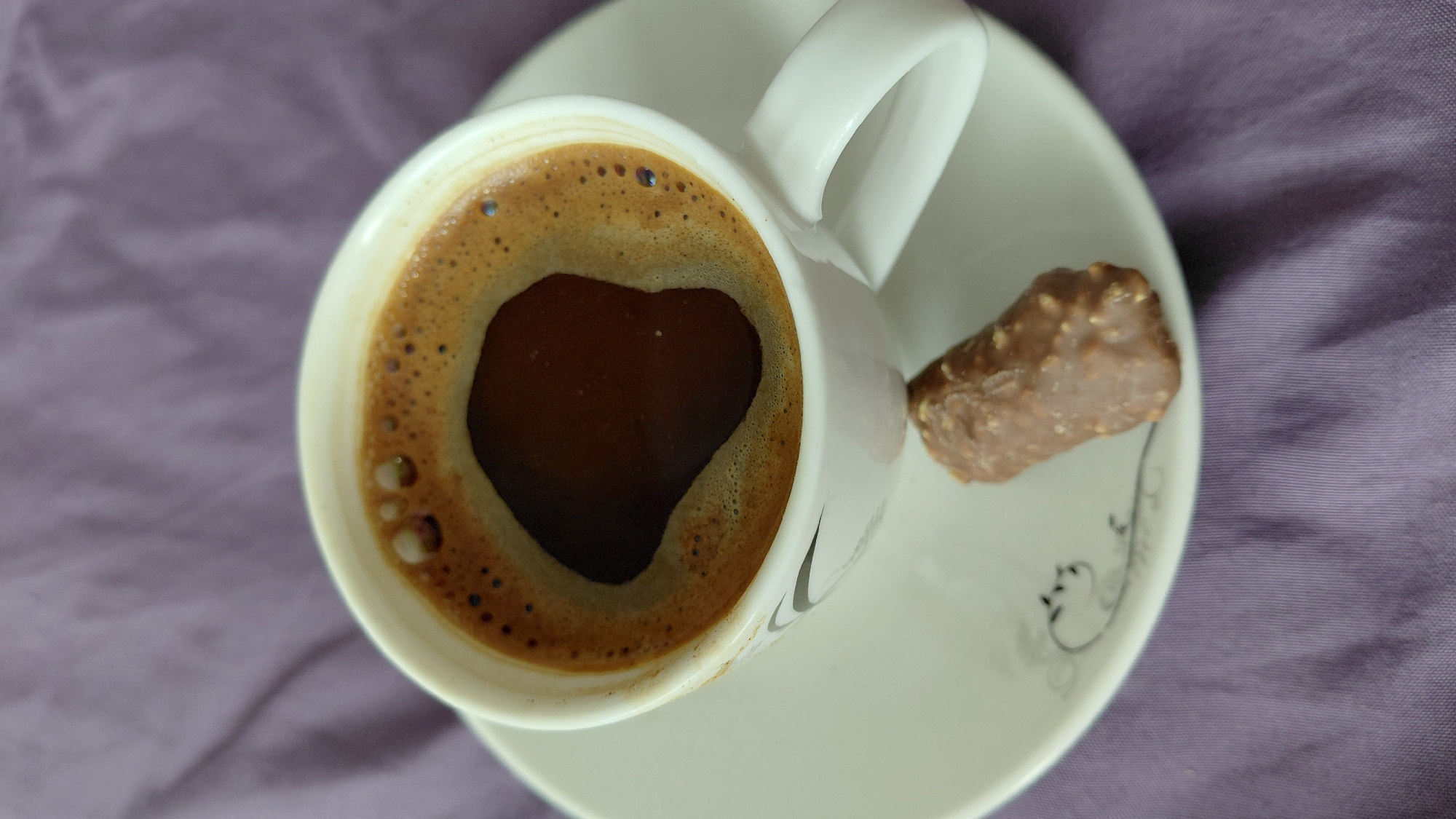 The image shows a close-up of a white cup of coffee with foam, sitting on a white saucer with a subtle gray swirl design. A chocolate-covered candy bar with nuts is placed on the saucer in front of the cup. The background is a soft, textured purple fabric, likely a tablecloth or bedspread. The scene is set in Abovyan, Armenia.
