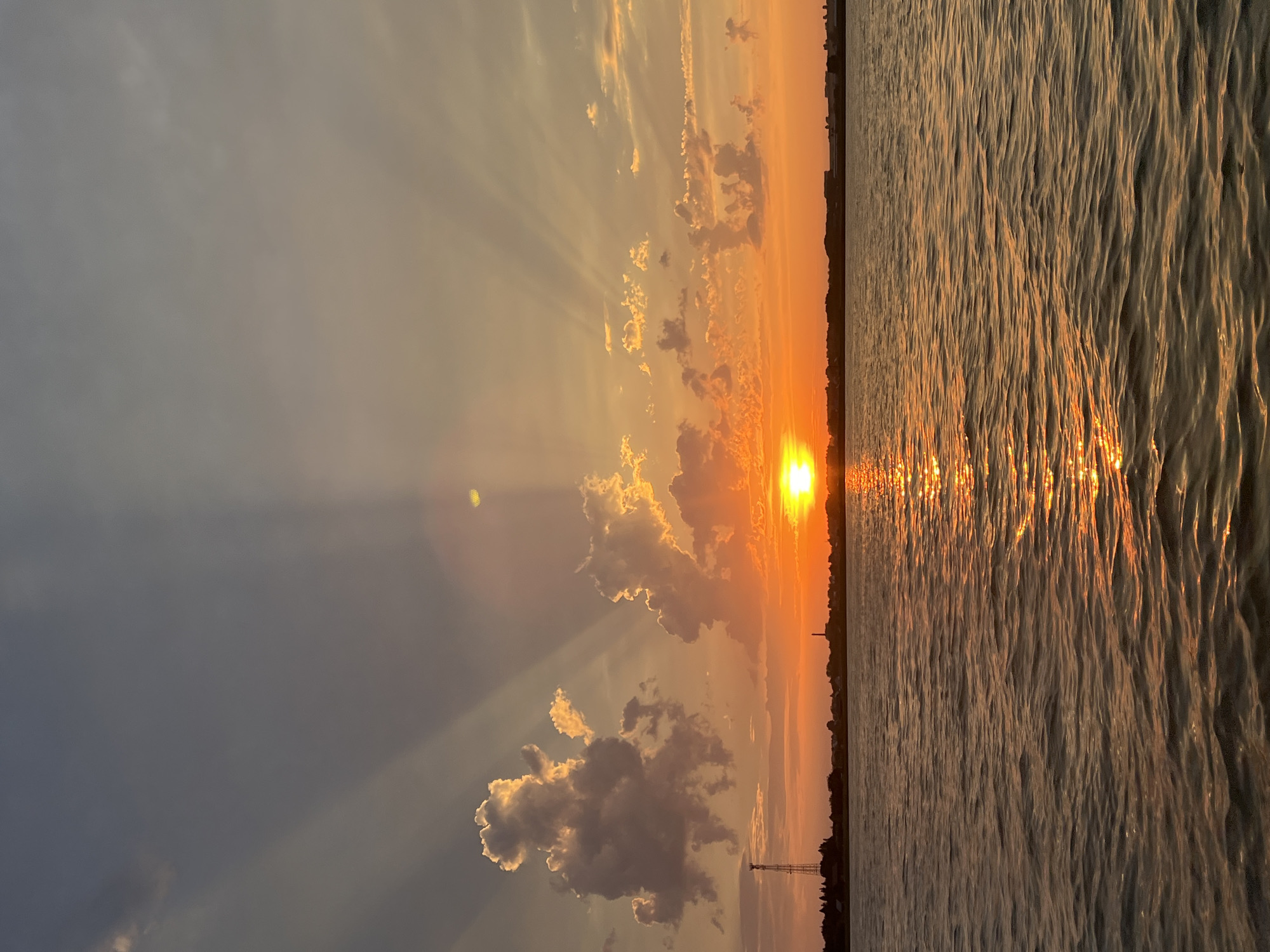 This image depicts a sunset over a body of water, likely a lake or river, in Novopichugovo, Russia. The sky is a gradient of orange and grey, with dramatic sunbeams radiating from the setting sun, which is a bright orange orb partially obscured by clouds. Wispy clouds, illuminated by the setting sun, are scattered across the sky. In the distance, a silhouetted shoreline is visible, featuring a few buildings and a tall, slender structure that appears to be a telecommunications tower. The water's surface is rippled, reflecting the warm hues of the sunset and creating a shimmering effect. There are no people or animals visible in the image. The overall scene is one of natural beauty and tranquility.