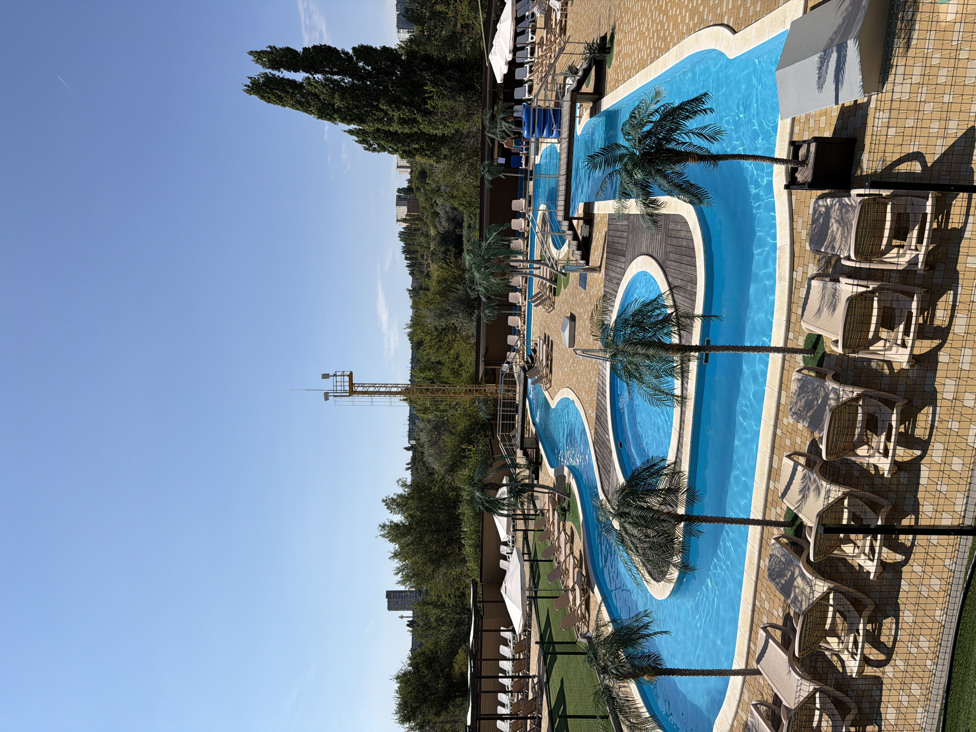 The image depicts a bright, sunny day at a water park located in Kamyshin, Russia. The park features a winding blue pool with a lazy river element, bordered by a light brown tiled deck. Several chaise lounges are arranged on the deck, some under white umbrellas, indicating a relaxation area for visitors. Palm trees, both real and artificial, are strategically placed around the pool and deck, adding to the tropical ambiance.
In the background, a yellow metal tower with lights and a platform stands tall, possibly a lifeguard tower or part of a ride. Beyond the immediate park area, dense green trees form a backdrop, with buildings visible in the distance, suggesting an urban or suburban setting. A faint contrail is visible in the clear blue sky. A person can be seen standing near the pool on a wooden walkway. The overall scene conveys a sense of leisure and outdoor recreation.