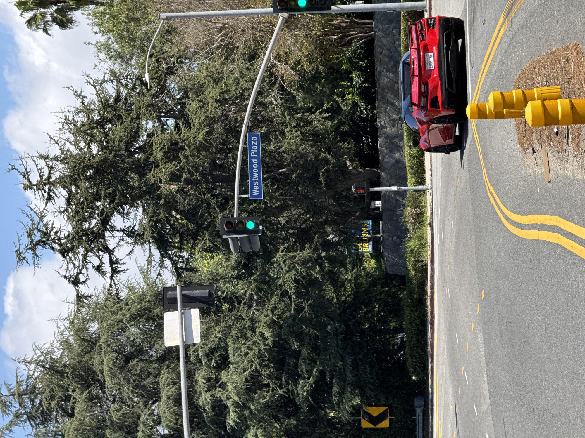 The photograph shows an intersection in the city of Los Angeles, California, United States. A street sign indicates the cross street is Westwood Plaza. A red Corvette with California license plate "LDS7 ME" is stopped at the intersection. The traffic light is green. There is a tree and foliage in the background. Yellow traffic barriers are along the side of the road.