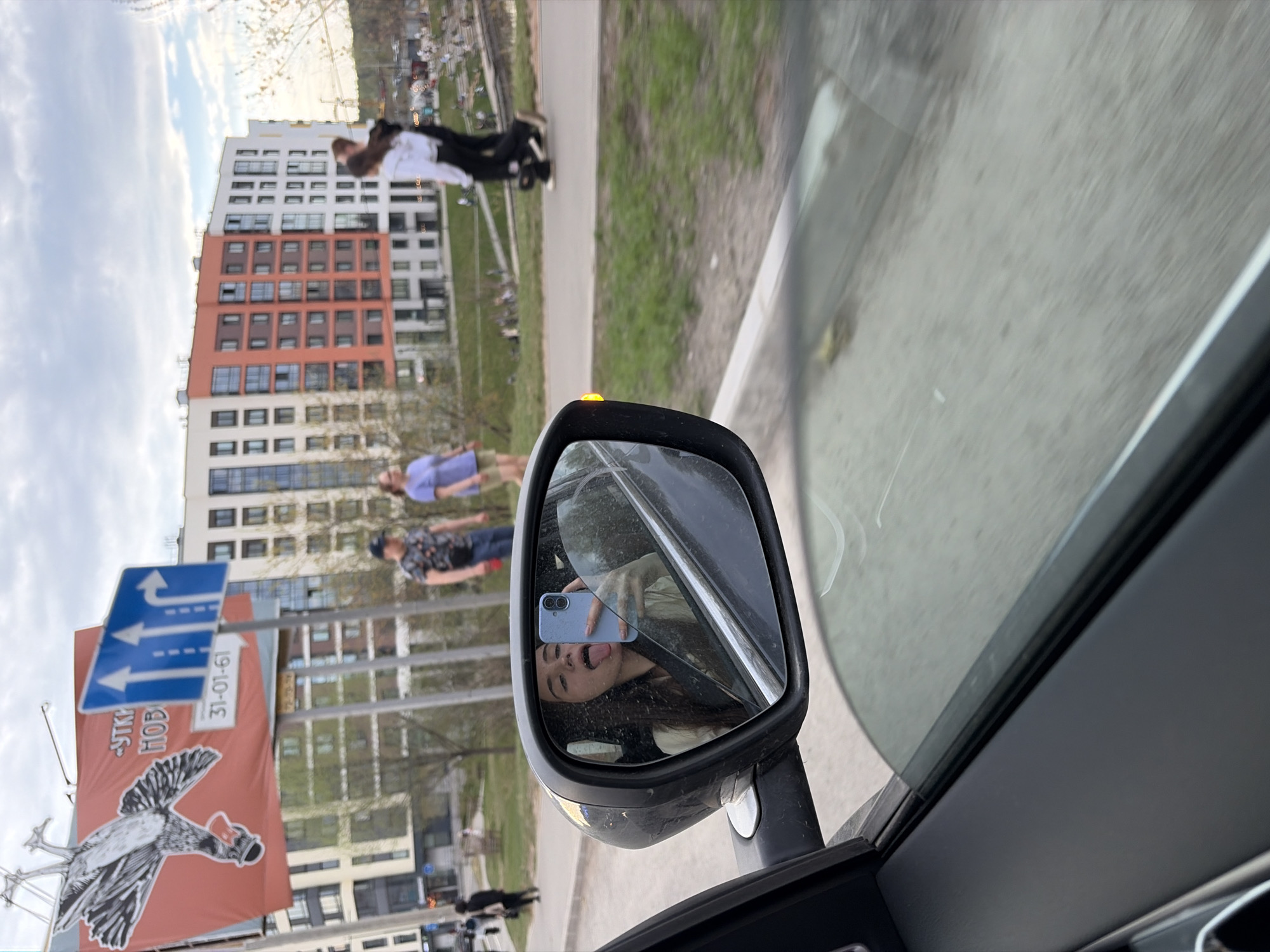 The image, taken from inside a car, shows a view of a street scene through the side mirror. In the reflection of the side mirror, a young woman is visible, holding a blue smartphone and sticking out her tongue. The street scene features several people, including a person pushing a stroller. There is a large banner depicting a bird wearing a hat and a sign indicating directions of traffic flow. A large building is present in the background. The banner is marked with "УТКА НОВО" (Duck New) and a date "31-01-61". The location is Izhevsk, Russia, based on the Cyrillic text on the banner. The car is driving on a road adjacent to a sidewalk and grassy area. The sky is overcast.