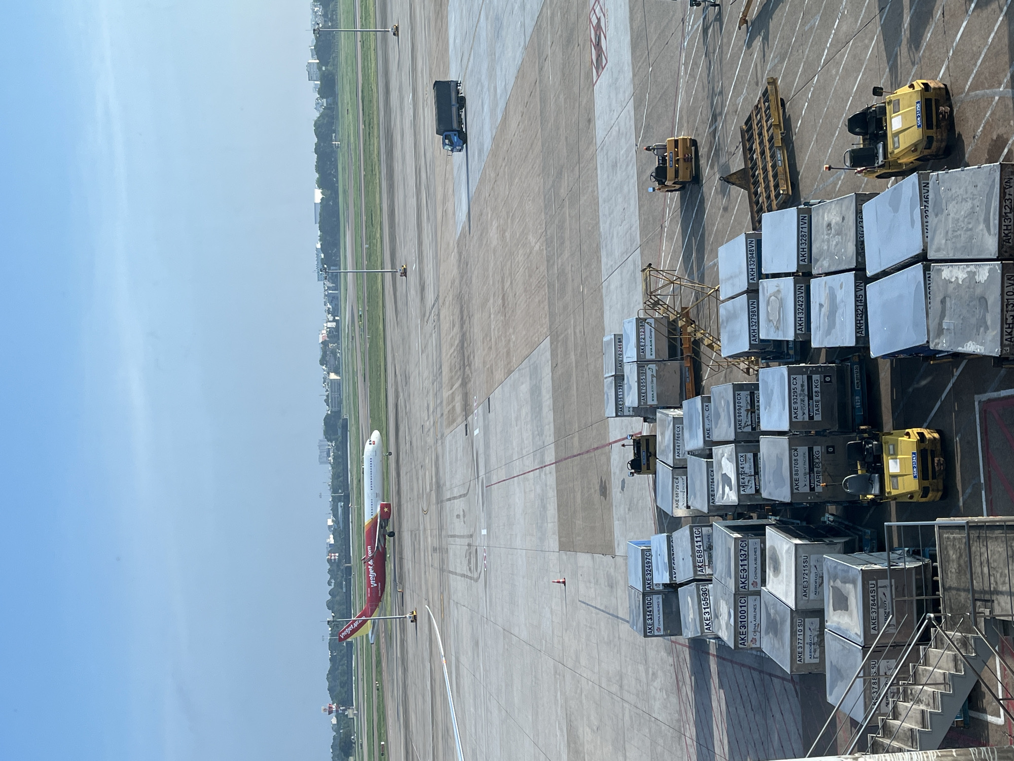 The image depicts an active airport tarmac under a clear, bright blue sky, most likely Tân Sơn Nhất International Airport (SGN) in Ho Chi Minh City, which serves the region including Thuận An, Vietnam. A red and white Vietjet Air Airbus aircraft, marked "Vietjet Air .com" on its fuselage, is positioned on a taxiway in the mid-ground, appearing to be taxiing or stationary.
In the foreground, numerous metallic Unit Load Devices (ULDs) or cargo containers are arranged on the concrete tarmac. Labels on these containers indicate various airline cargo operations, including "CHINA AIRLINES" (e.g., AKE 31410C, AKE 32497C, AKE 31001C, AKE 31137C), "AEROFLOT" (e.g., AKE 37710 SU, AKE 37215 SU, AKE 37844 SU), "CATHAY PACIFIC" (e.g., AKE 88729 CX, AKE 47766 CX, AKE 88764 CX, AKE 90930 CX), and "EVA AIR Cargo" (e.g., AKE 70551B, AKE 76246 BR, AKE 72123 BR). Some ULDs are also marked with "TARE 68 KG". Several yellow aircraft tugs are present; one in the immediate foreground shows the registration "SGN-31247", and another further back "SGN-31250", confirming the SGN (Tân Sơn Nhất) location. These tugs are connected to dollies carrying ULDs. A yellow flatbed cargo trailer is also visible. Metal stairs are partially visible on the left, leading up from the tarmac. An orange traffic cone stands on the tarmac near the center of the frame.
In the mid-ground, a blue box truck is driving across the tarmac. The background features extensive paved runways/taxiways, interspersed with strips of green grass. A distant control tower is visible to the left. The horizon reveals a dense line of trees, behind which an urban landscape of multi-story buildings extends, indicating a major city or metropolitan area. Utility poles with lights are distributed across the airport grounds. The overall scene suggests ongoing air traffic and cargo handling operations.
