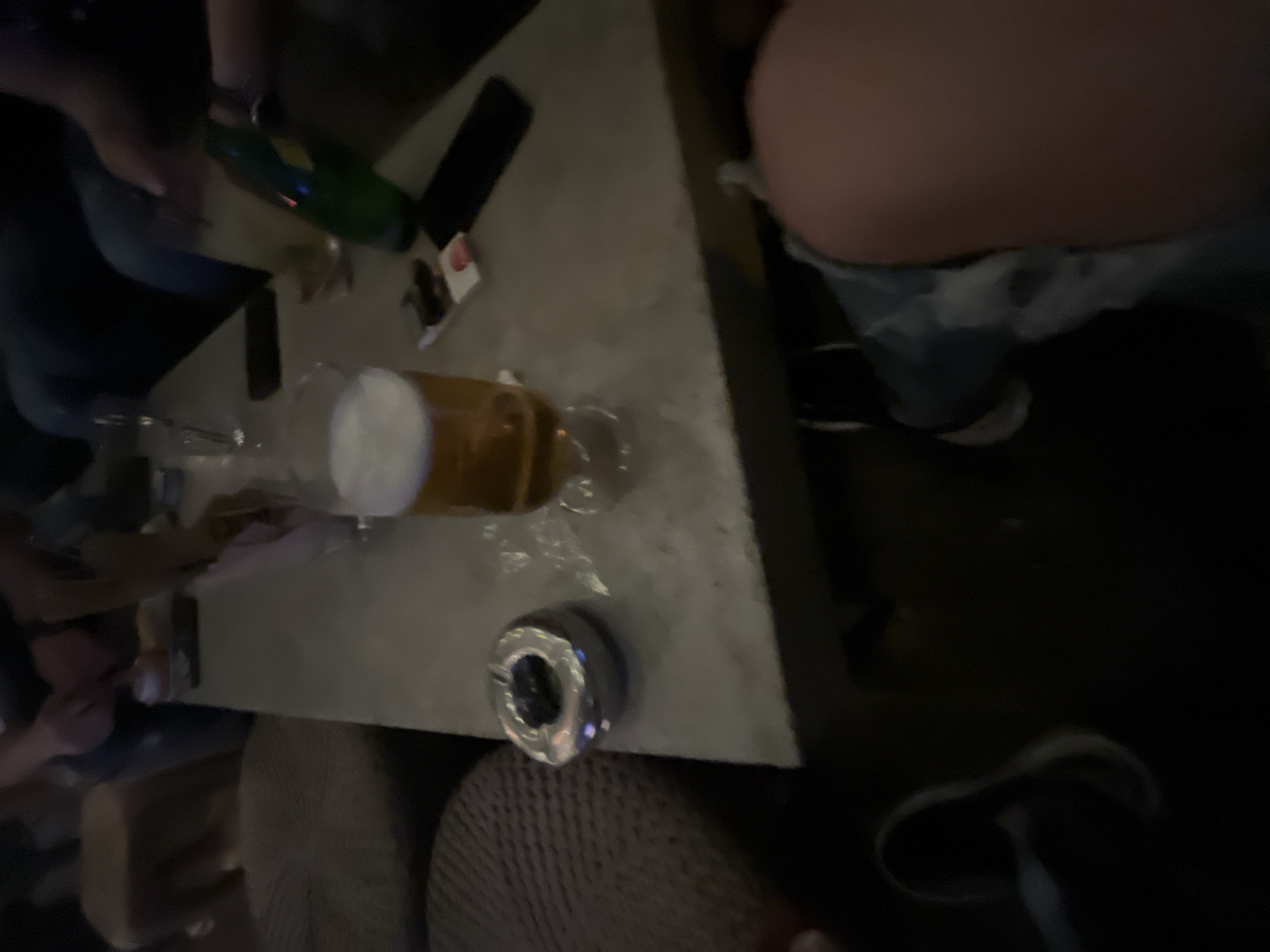 The image shows a cluttered table in a dark environment, possibly a bar or restaurant. A beer glass filled with beer sits prominently on the table. There is an ashtray and scattered items, including a bottle. The scene is dimly lit. The presence of an ashtray and beer suggests this may be a location with a focus on adult beverages and smoking. Location cannot be determined.