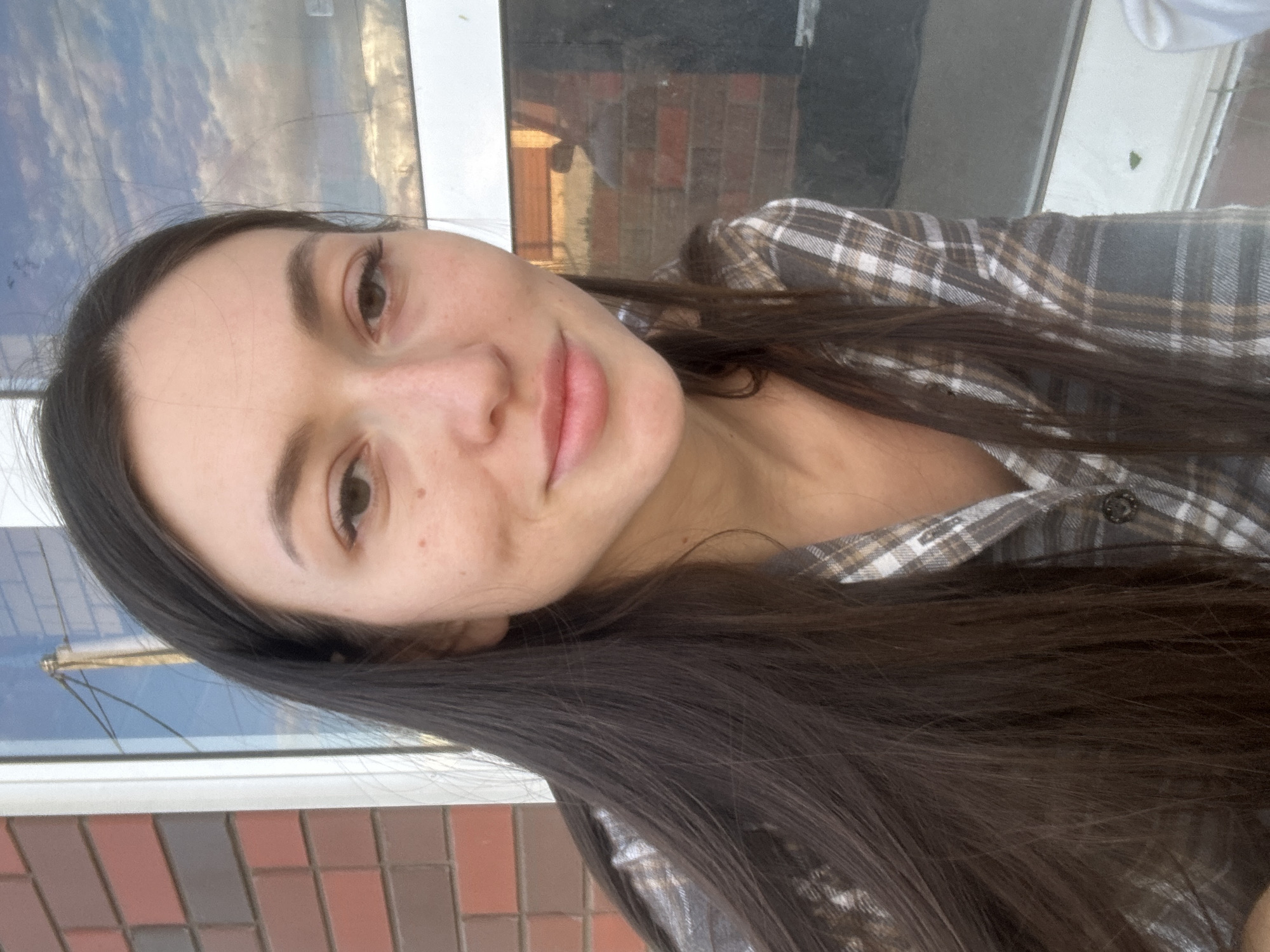 A young woman with long, dark brown hair is captured in a close-up, front-facing photograph. She is wearing a plaid shirt with brown, grey, and white patterns. Her face is tilted slightly to her left, and she has a subtle smile. The background features a brick wall on the left and a window with reflections of the sky and some structures. There is no identifiable location in the city of Эстачи, Russia.