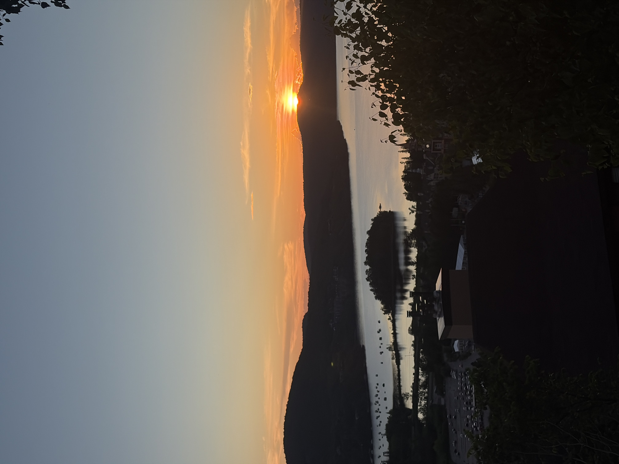 This photograph captures a serene sunset over a body of water, likely Lac Tremblant, with a mountainous backdrop. The sun, a bright orb of orange, is partially obscured by the silhouette of a distant hill, casting a warm glow across the sky. The sky transitions from a soft blue at the top to hues of orange and yellow near the horizon, with wispy clouds tinged pink by the setting sun.
In the foreground, dark silhouettes of trees frame the scene, their leaves creating a textured pattern against the lighter sky. Below, a tranquil lake reflects the twilight colors. Dotted across the water are numerous small boats, their forms indistinct in the fading light.
Further in the midground, a small island with trees rises from the lake. On the shore, a collection of buildings, possibly part of the Mont-Tremblant resort area, are visible. These structures are silhouetted against the water and sky, with some architectural details, such as pointed roofs and chimneys, discernible on closer inspection. Adjacent to these buildings, a parking lot filled with cars can be seen, suggesting a populated area.
The overall impression is one of natural beauty and peacefulness, characteristic of a lakeside retreat in Mont-Tremblant, Canada, during sunset.