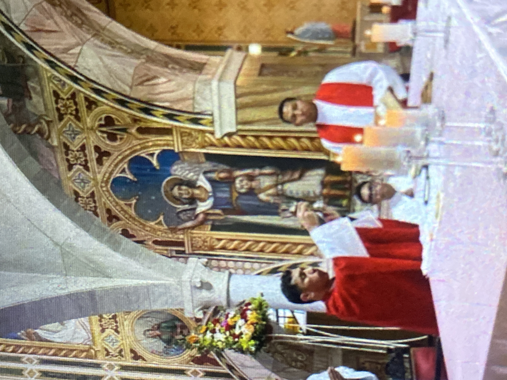 The image shows the interior of a church. Three men, possibly priests, are present. One man in a red vestment is at the forefront, holding what appears to be a small white object. Another man in a red and white vestment stands behind him, while a third man in white stands further back. On the altar, there are lighted candles and glassware. The church's architecture features arches, paintings, and detailed ornamentation. An icon of what appears to be a religious figure is visible on the wall. Floral arrangements can also be seen. The location is Tinaquillo, Venezuela.
