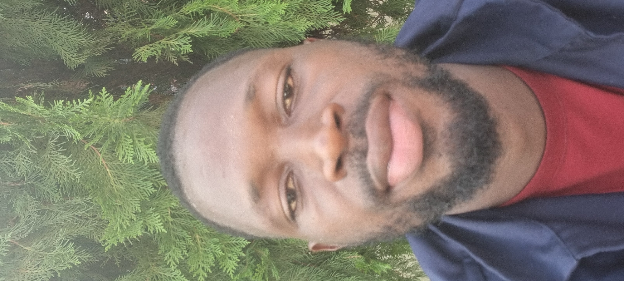 A close-up, front-facing selfie shows a dark-skinned Black male looking directly at the camera. He has a beard and is wearing a dark blue jacket over a red t-shirt. The background is dominated by green foliage. The image is undated and unlocated, but if placed in Moba, Nigeria, it would depict an outdoor scene, possibly in a garden or park, on a moderately lit day. There are no visible actions or interactions, and the subject's expression is neutral. The environment appears natural and undeveloped.
