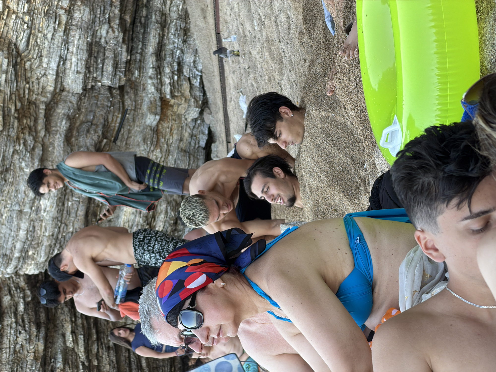 The image depicts a beach scene in Budva, Montenegro, characterized by a rocky cliff face serving as a backdrop. Several individuals are present, engaged in various activities. In the foreground, a woman wearing sunglasses and a blue bikini top with a colorful headscarf is seated, facing left. Beside her, a young man is lying down, his head resting on a scarf. Further back, two young men are partially buried in the sand, with only their heads visible. Another man, shirtless and wearing patterned shorts, stands to the right, looking at his phone. To the left, a young woman in a blue top is also visible. Objects in the scene include water bottles, a phone, and a bright green inflatable ring. The overall atmosphere suggests a casual gathering on a sunny day at the beach.