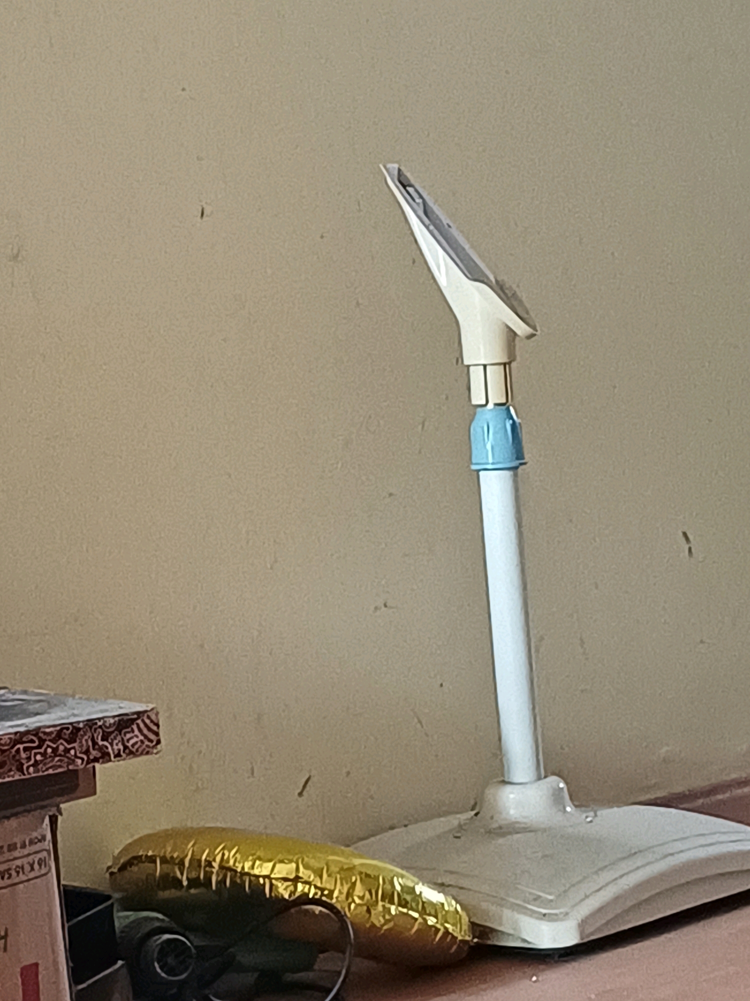 The image shows a floor fan with a white base and pole, a blue connector, and a white fan head. The fan is positioned on a tiled floor, with a beige wall in the background. To the left of the fan, there is a stack of boxes. A shiny, golden object, possibly a balloon or wrapping paper, lies on the floor near the base of the fan. The scene appears to be indoors. The location is not specified in the image.