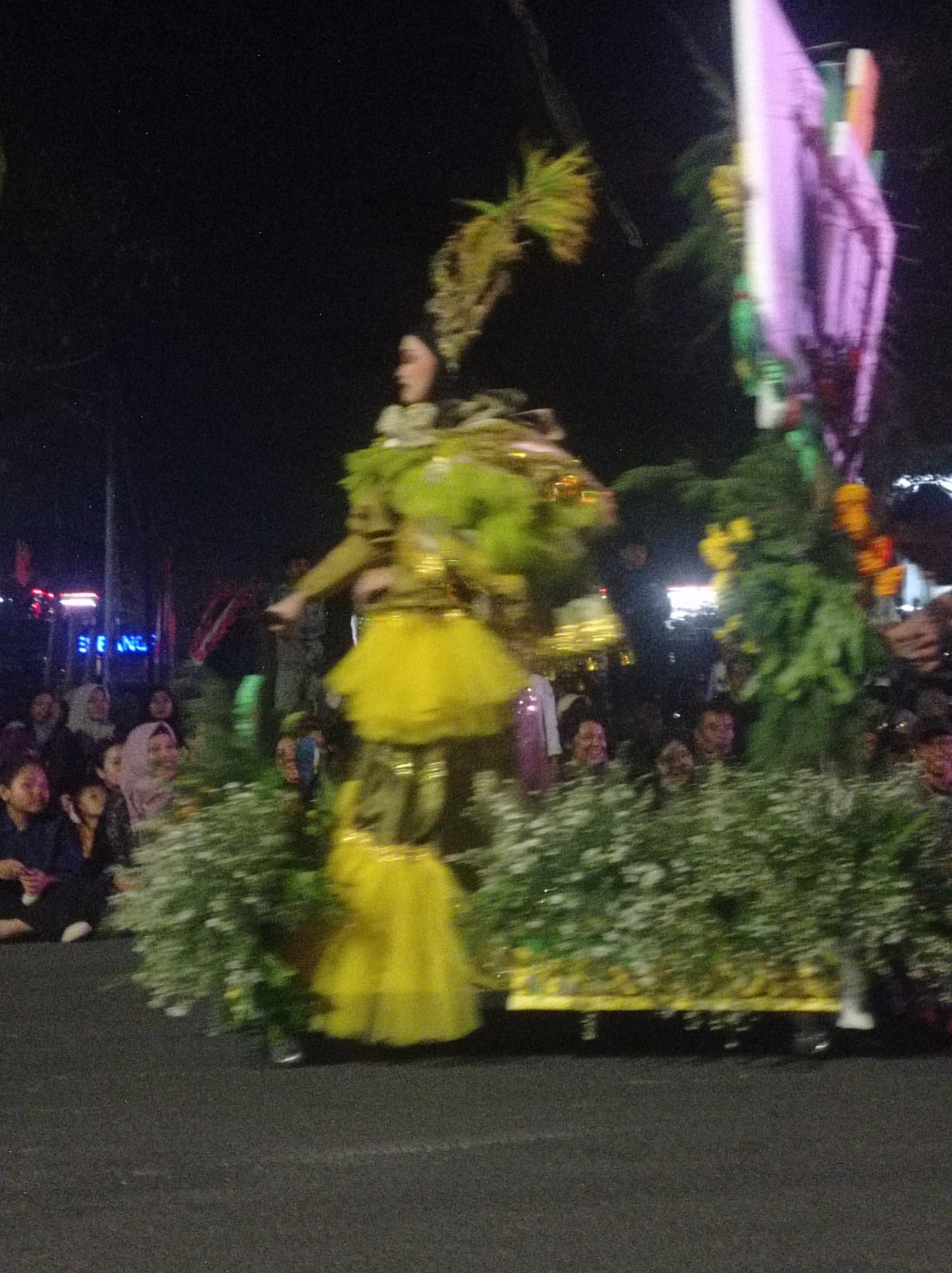 The image shows a blurry night scene, likely a parade or festival, in the city of Pandean, Indonesia. A person in a vibrant yellow costume with a green, leafy headdress is the central figure. They are on a float adorned with greenery and what appear to be floral decorations. In the background, a crowd of people are watching the event, their faces partially visible and their expressions suggesting engagement with the spectacle. The environment is dark, illuminated by artificial lights from the float and possibly streetlights, creating a festive atmosphere.