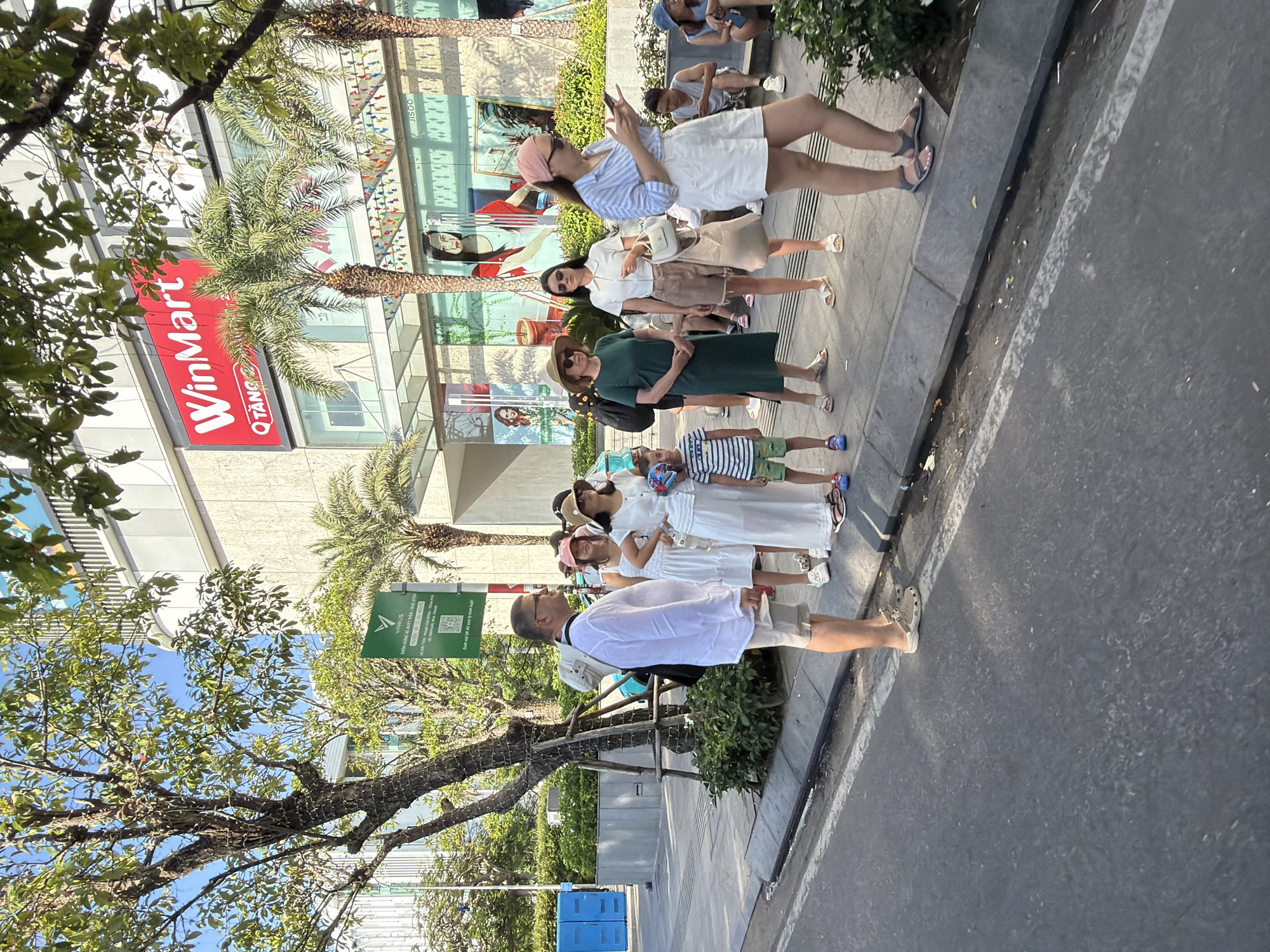 The image depicts a sunny outdoor scene in Nha Trang, Vietnam, specifically in front of a WinMart store. A group of people, including adults and children, are standing on a sidewalk next to a street. The architecture suggests a modern urban environment, with a large building displaying the "WinMart" logo. Palm trees and other greenery are visible, indicating a tropical climate. The people are dressed in casual summer attire. Some individuals are looking at their phones, while others are engaged in conversation or observing their surroundings. The atmosphere appears relaxed and leisurely.