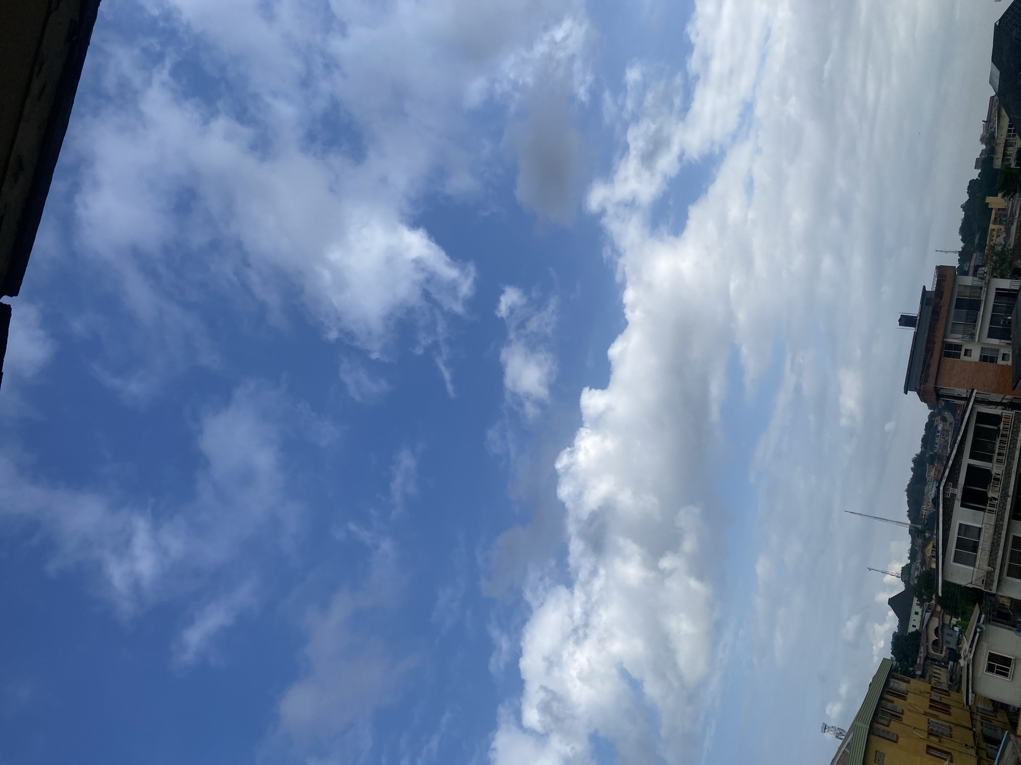 The image shows a view of the sky over Abeokuta, Nigeria. The sky is filled with blue and white clouds. Below the sky, there are several buildings, some of which are multi-story. The buildings appear to be a mix of residential and commercial properties. There are also some trees and a hill in the background. The overall impression is of a moderately populated urban or semi-urban environment on a cloudy day.