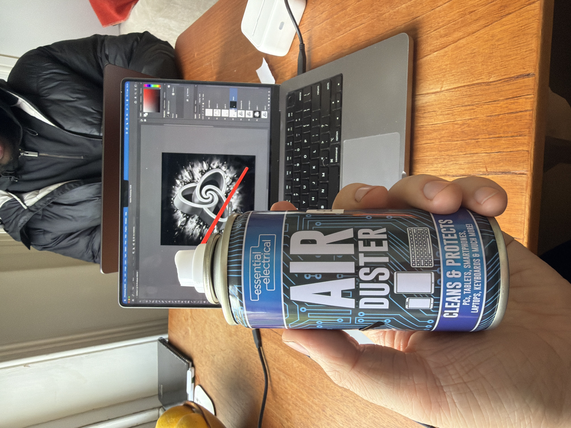 A person is holding a can of "Essential Electrical Air Duster" in front of an open laptop displaying an image being edited in Adobe Photoshop. The laptop rests on a wooden desk. A white charging block is positioned near the laptop. Part of a person wearing a black jacket is visible to the left of the laptop. A black cable is attached to the side of the laptop. The location is not discernible from the given image.