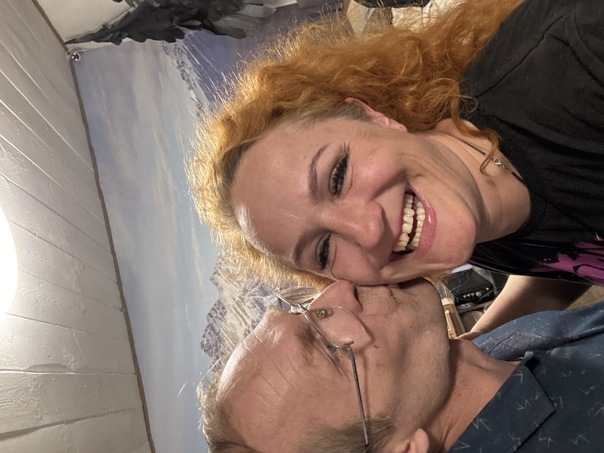 A selfie captures a Caucasian woman with curly red hair, wearing a black shirt and a silver necklace, smiling broadly at the camera. She is positioned slightly above and to the right of a Caucasian man with short, light hair and glasses. The man is wearing a blue shirt with a pattern. The background features a wall with what appears to be a mural of a mountain range and a bird figure at the top. A white wall with horizontal paneling is visible to the left. The photograph provides no contextual information to indicate a specific location in the city of Chaikovsky, Russia, nor any details that would definitively associate the scene with this city.