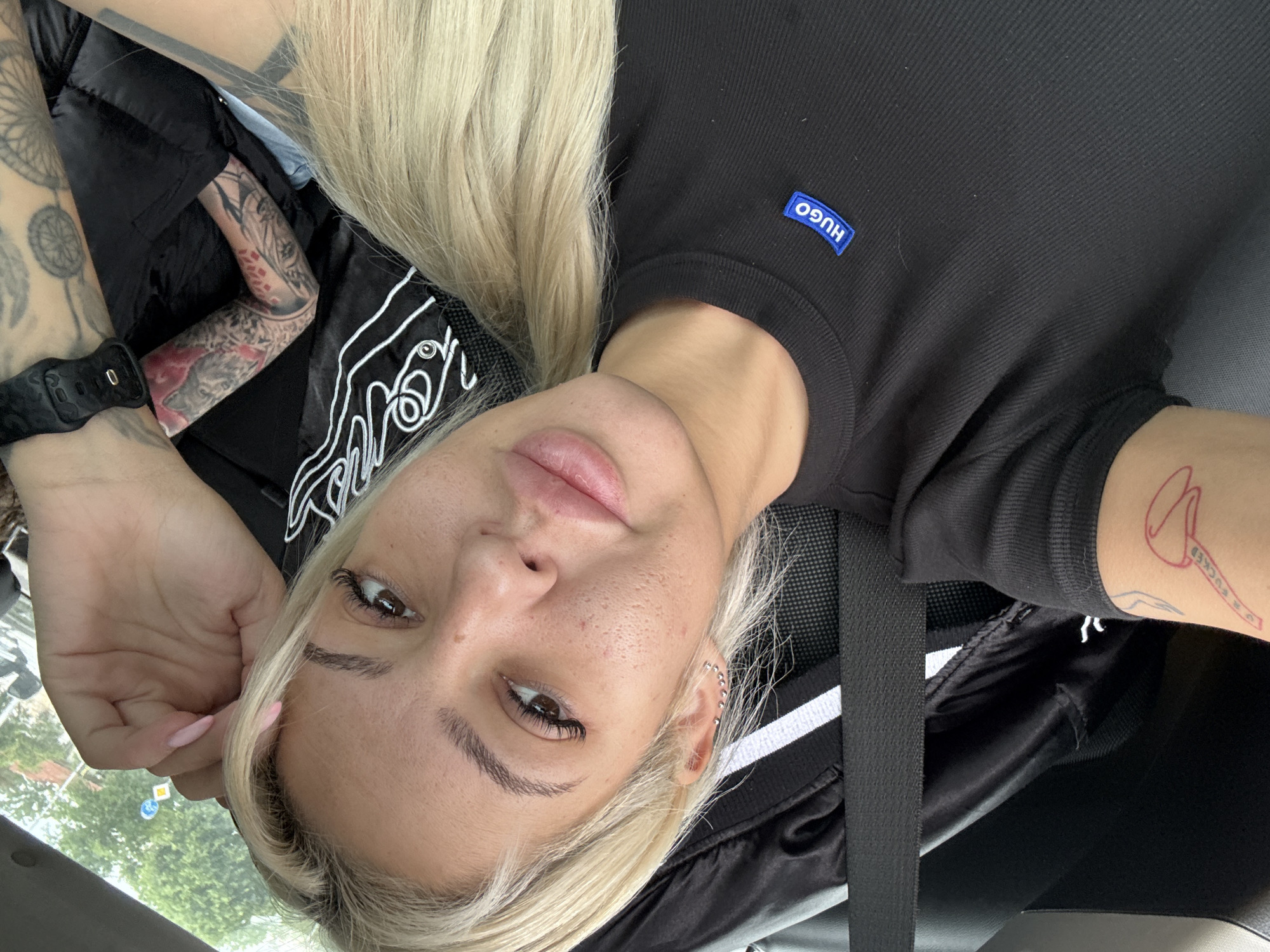 A medium shot depicts a blonde-haired woman inside a vehicle. She is angled to the camera, with a black Hugo brand t-shirt visible. She is wearing a black seatbelt. The woman has multiple tattoos on her arms. The setting appears to be daytime. A partial view of exterior scenery is visible. The location in the city of Plovdiv, Bulgaria is not discernible.