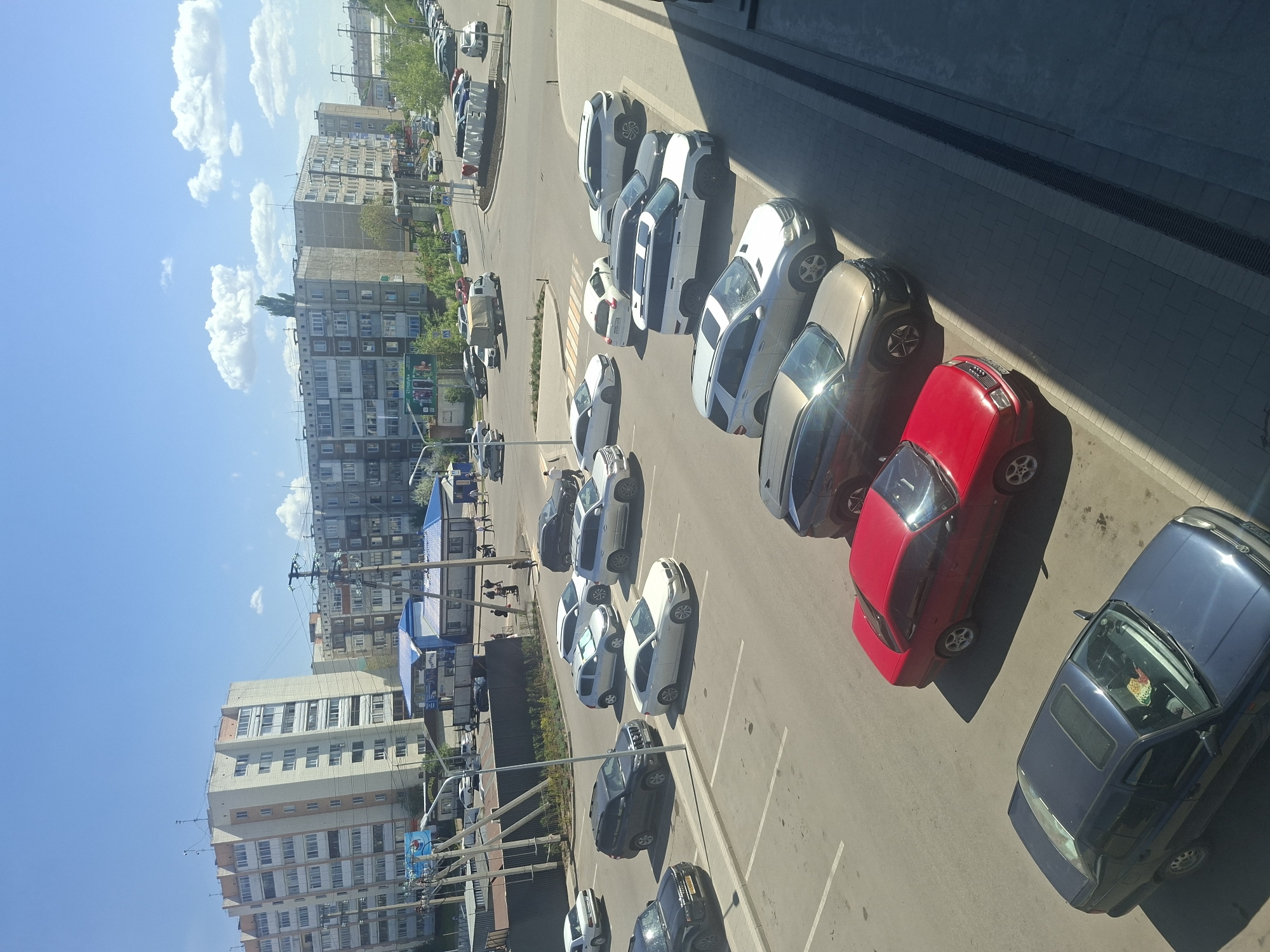 This is a high-angle shot, seemingly taken from an elevated position, showing a parking area near a building in Kokshetau, Kazakhstan. The area is urban, with multi-story residential buildings visible in the background under a partly cloudy sky.
Several cars of varying colors (white, gray, black, red, and blue) are parked in designated spaces. The parking area appears to be adjacent to a sidewalk and a street with pedestrian activity. The presence of power lines and streetlights suggests a typical urban infrastructure. The lighting suggests a sunny day.