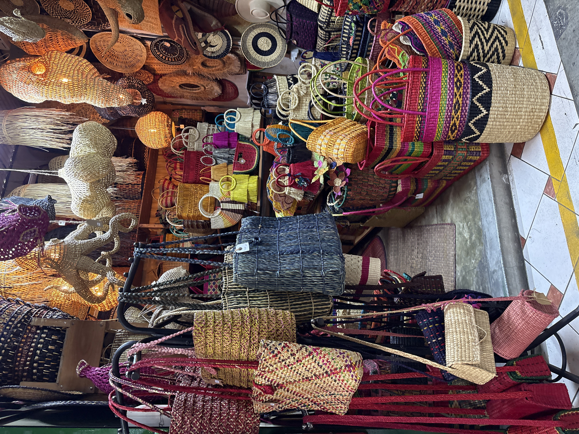 This image depicts a vibrant stall in Miraflores, Peru, filled with an extensive assortment of handcrafted woven items. The stall is densely packed with various types of bags, baskets, and decorative hanging light fixtures, all made from natural, woven materials such as straw and reeds.
In the foreground, a rack displays an array of colorful tote bags and shoulder bags. These range in size and design, from intricately woven patterns in reds, yellows, and blues, to more natural straw-colored bags. One prominent bag in the lower right is a large, cylindrical woven basket with a colorful striped pattern of magenta, green, yellow, and black, stacked on top of another basket with a black and beige zigzag design.
The background is filled with an abundance of hanging light fixtures shaped like marine life, such as octopuses, and abstract organic forms, emitting a warm, inviting glow. These are interspersed with woven wall hangings and larger baskets. The overall impression is one of a bustling marketplace offering artisanal crafts.
The floor is tiled with a checkered pattern of white and reddish-brown squares, accented by yellow painted lines, suggesting an outdoor or semi-covered market setting. No people are visible in the image, focusing the viewer's attention entirely on the products and the ambiance of the stall. The lighting is a mix of ambient market light and the warm glow from the woven lamps.