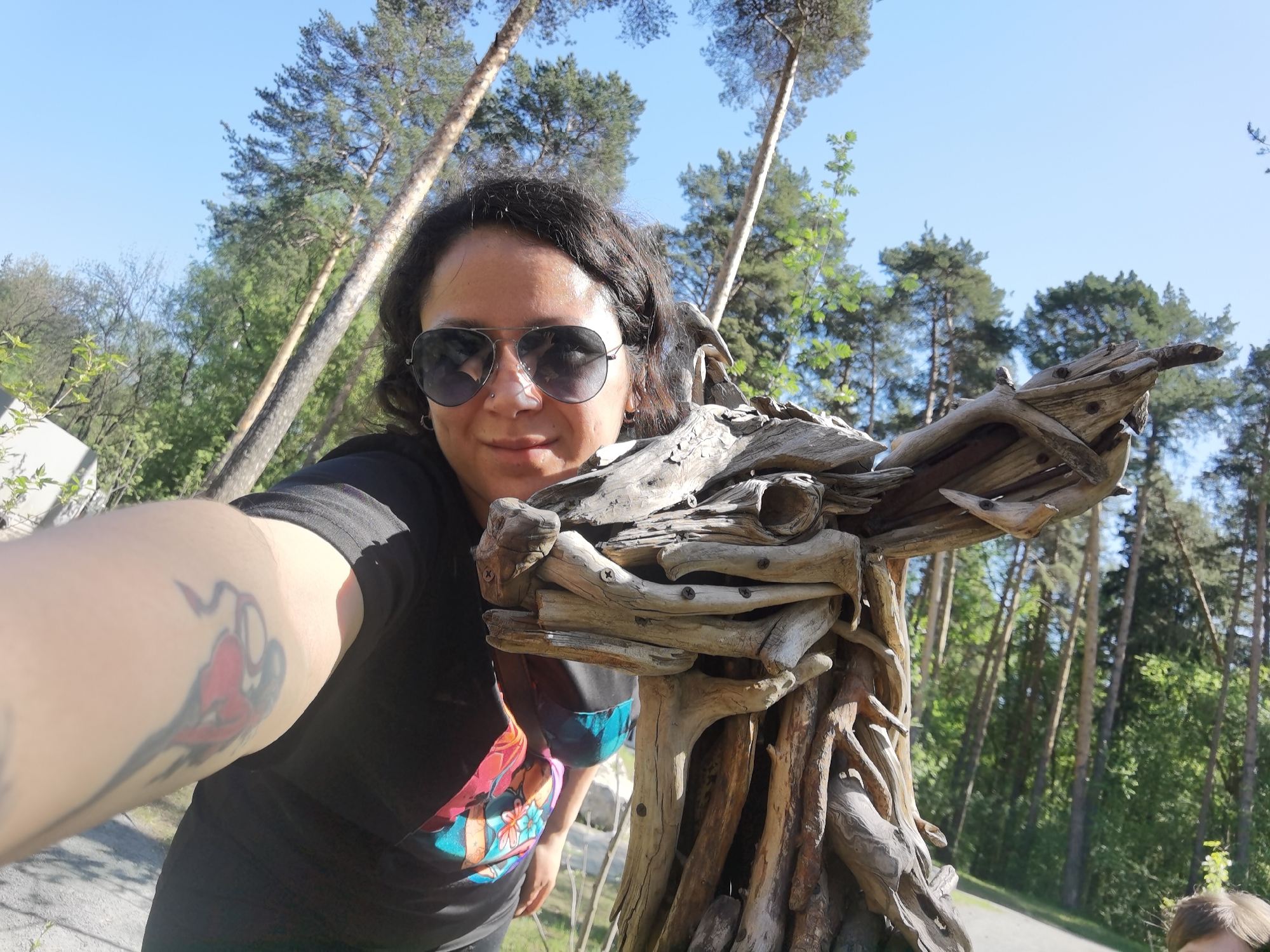 A person takes a selfie with a wooden sculpture in Perm, Russia. The person has dark hair, sunglasses, and a tattoo on their left arm, wearing a dark t-shirt. The sculpture, made of driftwood, appears to be a stylized animal head. The background includes trees and a path, suggesting an outdoor park or forest setting. The sky is blue, indicating a sunny day.