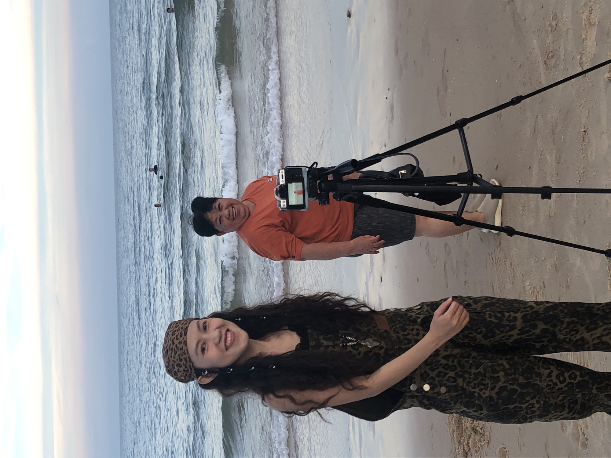 Two women are standing on a sandy beach in Phu Thuy Ward, Vietnam, with the ocean behind them. The woman on the left is younger, with long dark curly hair and is wearing a leopard-print bandana and a matching jumpsuit. She is smiling at the camera. The woman on the right is older and is wearing an orange t-shirt and grey shorts. She is also smiling. A camera is mounted on a tripod between them, facing the older woman, and it appears to be recording. In the background, several people are visible swimming and wading in the ocean. The sky above is a pale, soft color, indicating either dawn or dusk.