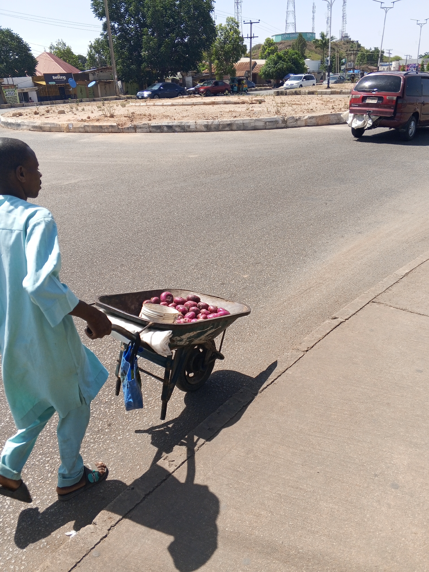 A man walks beside a road, pulling a wheelbarrow filled with red onions. He is wearing a light blue tunic and sandals. The wheelbarrow also holds a white container and a blue plastic bag. The location is Jos, Nigeria, based on landmarks in the background. Behind the man, a red van is driving on the road, and there are other cars visible in the distance. A roundabout with sparse vegetation is also visible. Power lines and communication towers can be seen on a hill in the background.