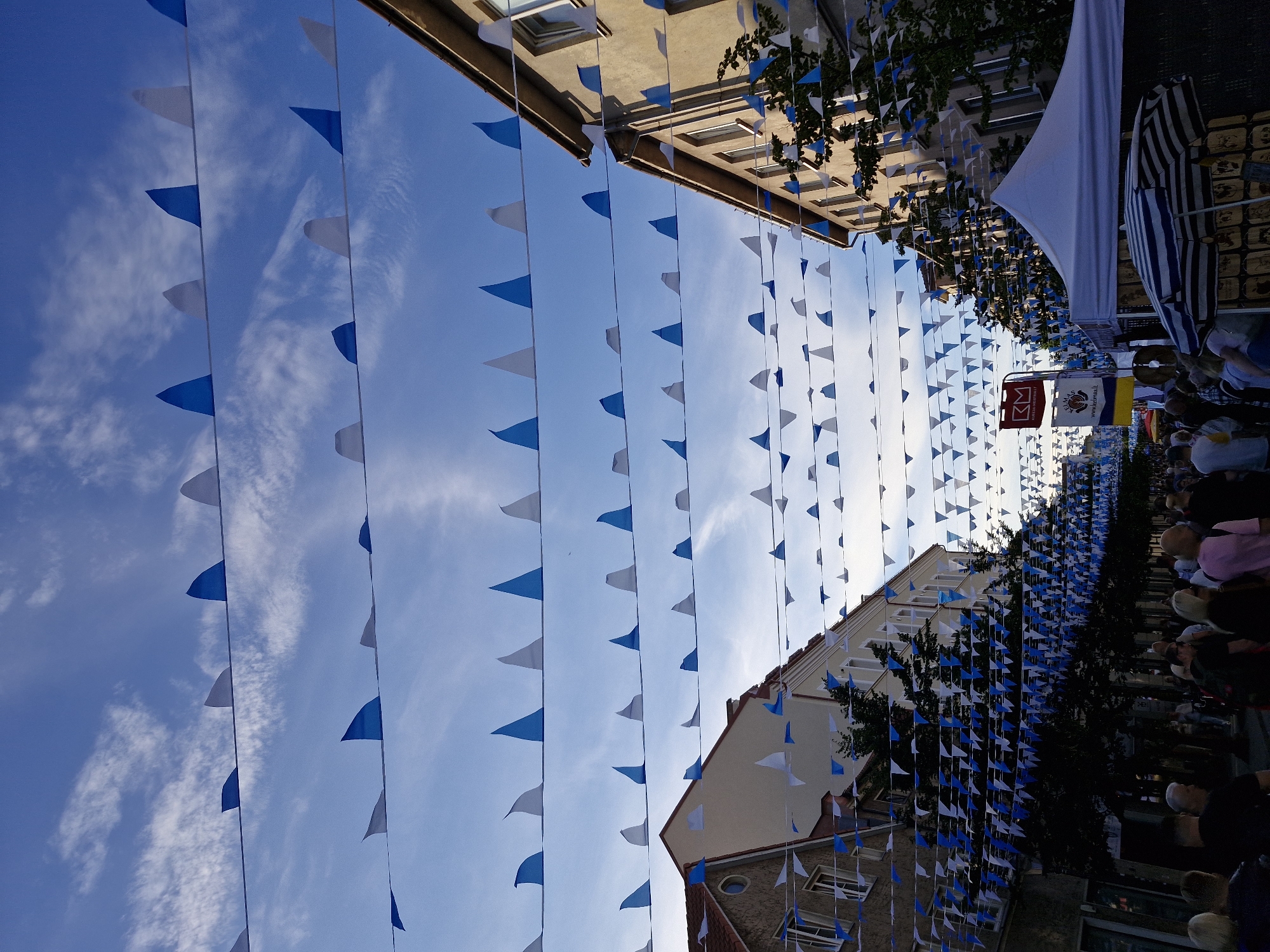 The image depicts a street scene in Klaipėda, Lithuania, likely during a festival or event. Multiple rows of triangular blue and white flags are strung across the street, creating a festive atmosphere. The flags are suspended above a crowd of people walking along the street, suggesting a public gathering. On either side of the street, there are buildings with a traditional European architectural style, characterized by their multi-story design and visible windows. Some of these buildings appear to be shops or commercial establishments, indicated by the presence of awnings and what seem to be market stalls. A white tent is visible on the right side of the image, possibly a vendor's booth. In the background, a red sign with the letters "RM" is displayed. The sky is blue with scattered white clouds, suggesting daytime. The overall impression is one of a lively street event taking place in a city setting.