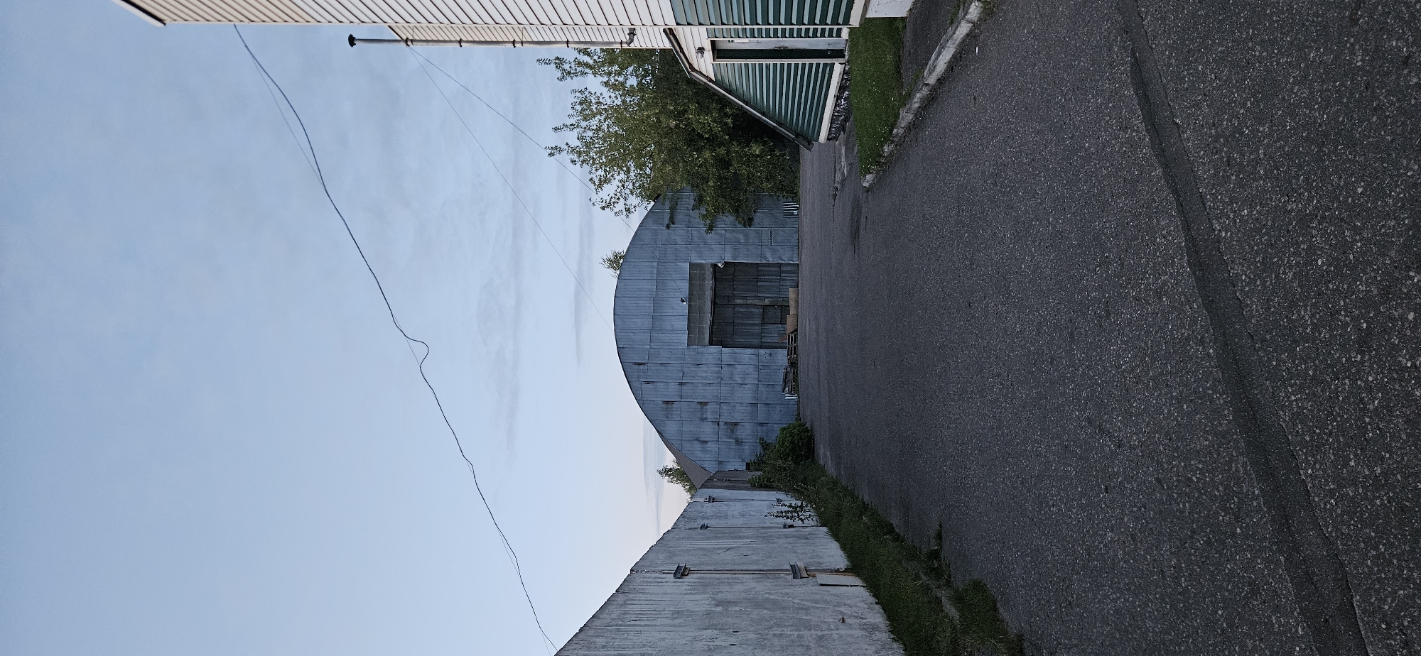 The image depicts an outdoor industrial or storage area. In the foreground, a paved asphalt road leads toward a large, arched metal building with double doors. To the left of the road is a tall, grey concrete wall with visible vertical seams. To the right, a smaller, light-colored building with green siding and a sloped roof is visible. Trees and foliage are present behind the main metal building. The sky is a pale blue with wispy clouds. A thin wire stretches across the sky. There are no people visible in the image. The scene appears to be located in an industrial zone, possibly on the outskirts of Vitsebsk, Belarus.