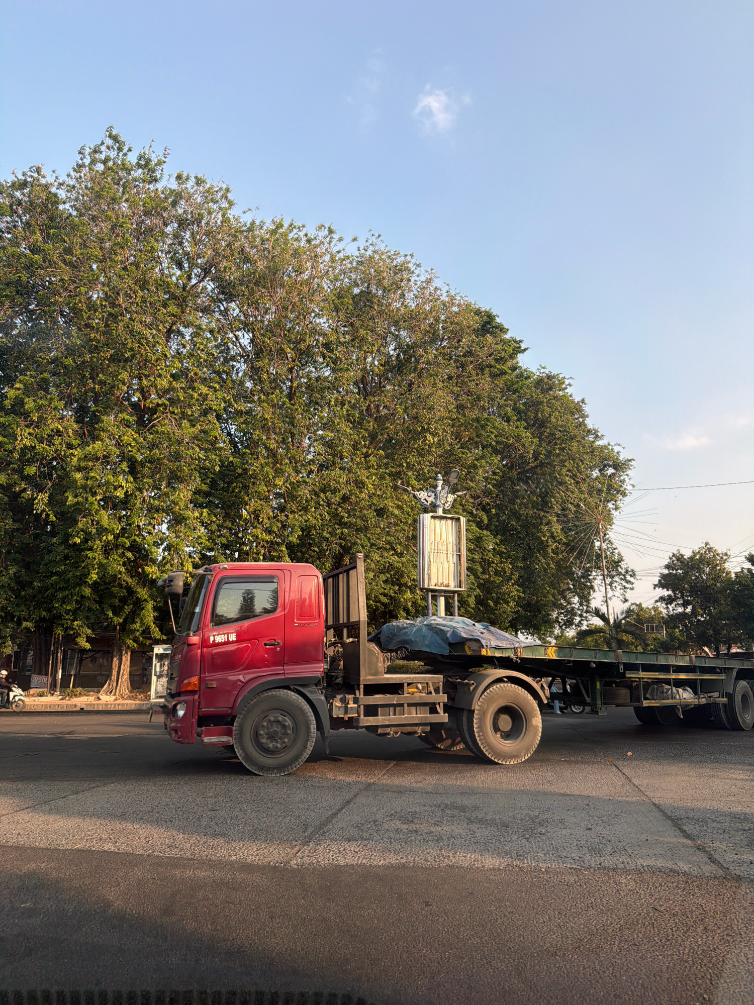 In Talkandang, Indonesia, a red flatbed truck is parked on a paved road in front of a dense line of large trees. The truck has its trailer attached and covered with a blue tarp. The sky above is clear with a few wispy clouds. The sun appears to be low in the sky, casting long shadows and illuminating the scene with a warm light. The surrounding environment suggests a daytime setting, likely late afternoon or early morning, given the lighting.