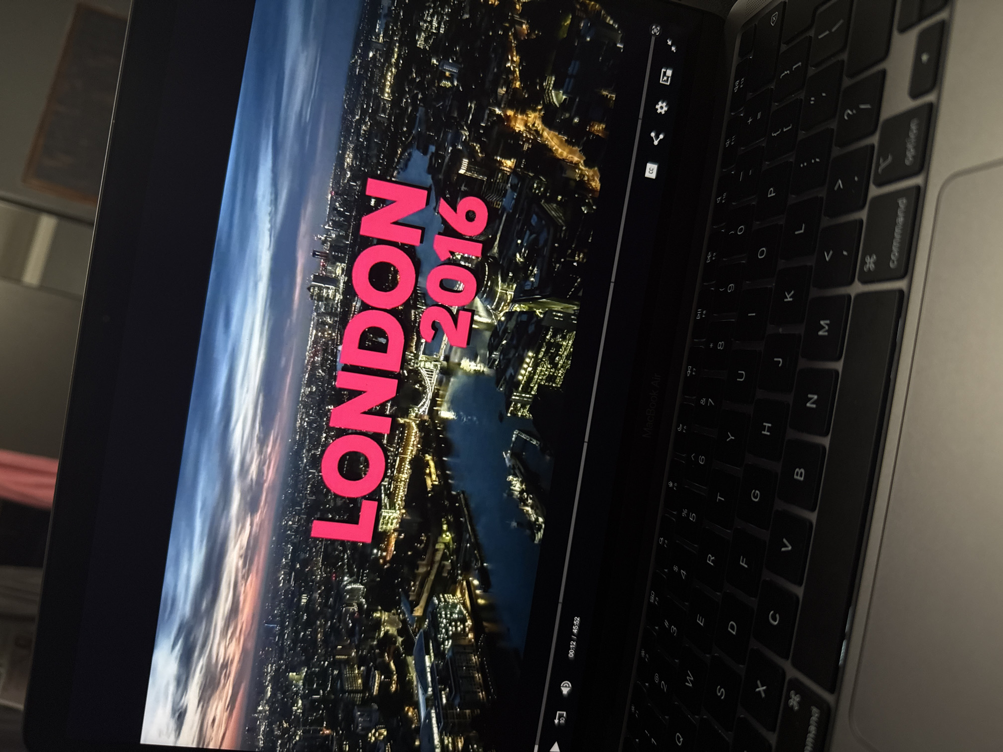 The image shows a laptop displaying a scene of London at night, with the text "LONDON 2016" overlaid in pink. The aerial view of London shows a river cutting through the city, which is illuminated by streetlights and building lights. To the right of the screen, parts of the laptop's keyboard are visible, including the keys for letters, numbers, and function keys like "command" and "option". The laptop is situated in an indoor environment, where the lighting seems to be subdued.