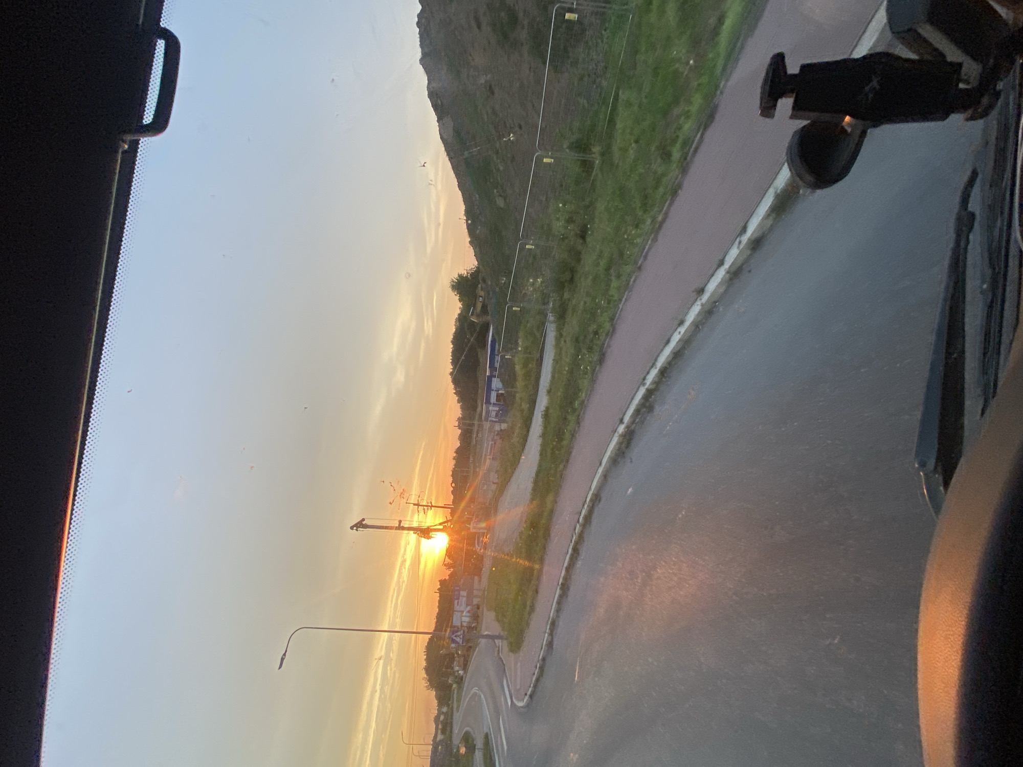 The image captures a sunset over a construction site, as viewed from inside a vehicle. The sun is low on the horizon, casting a warm glow across the sky and illuminating a construction crane and power lines. A road curves to the left, with a sidewalk and grassy embankment running alongside it. The embankment is fenced off with temporary barriers. The foreground shows a portion of the vehicle's dashboard and windshield wipers. There are no people visible in the image, and the focus is on the landscape and the construction activity. The scene appears to be in Podłęże, Poland, given the context.