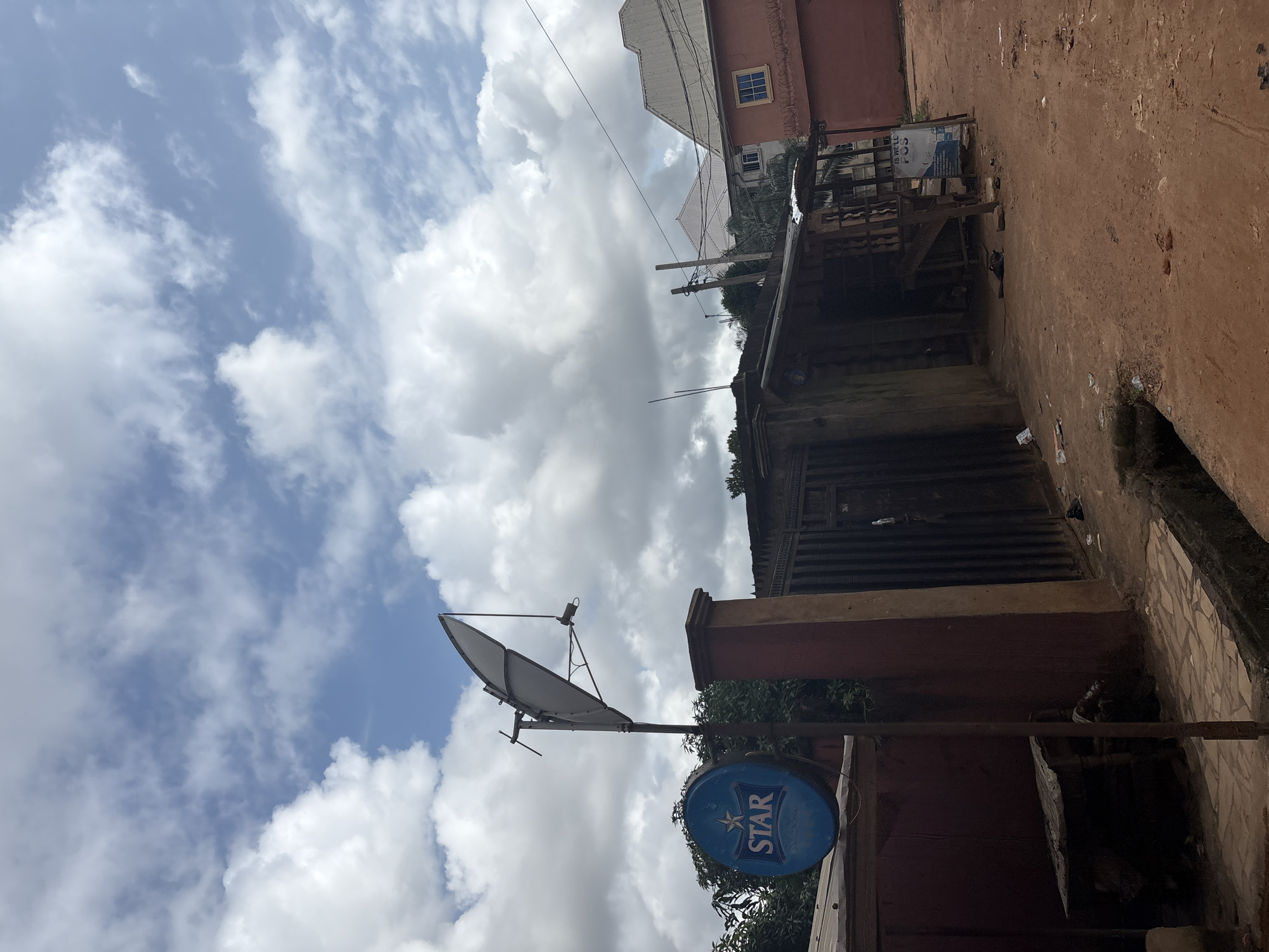 The image depicts an outdoor scene in Nkpor, Nigeria, featuring a satellite dish mounted on a pole, a blue circular sign with the word "STAR" prominently displayed, and a building with a metal gate. The ground is unpaved and appears to be made of dirt and some scattered debris. In the background, there are other buildings, trees, and a cloudy sky. The overall environment suggests a modest urban or semi-urban setting.