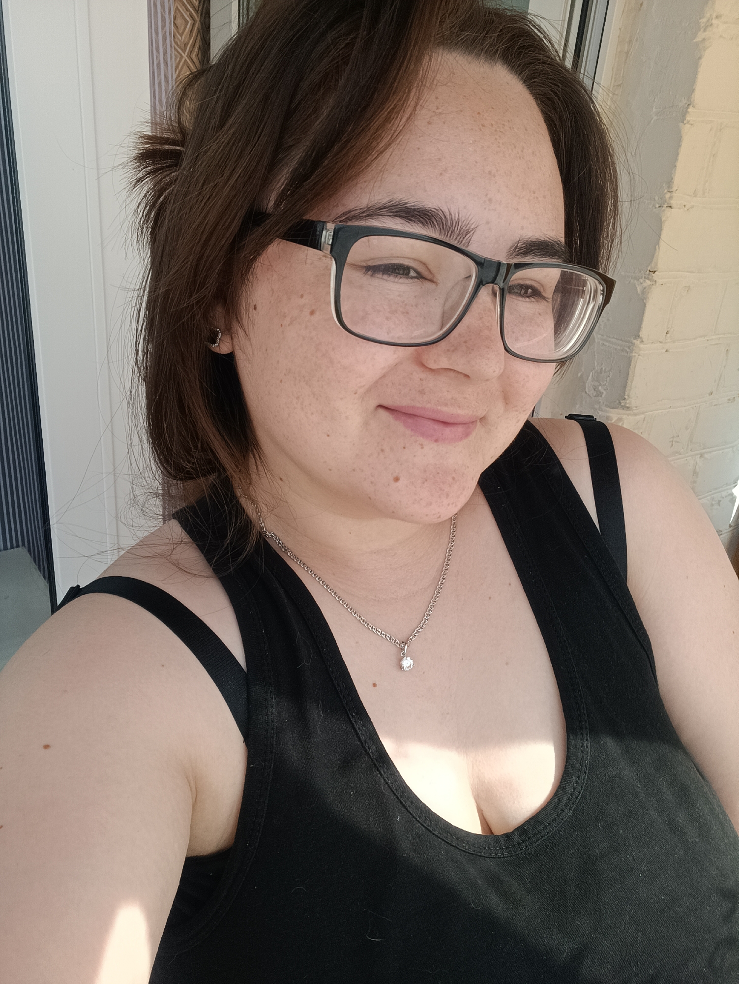 The image is a selfie taken outdoors, showing a woman with brown hair and fair skin with freckles. She is wearing glasses with black frames and a black tank top, with a silver necklace and a pendant. The background shows a brick wall and what appears to be vertical blinds. There is no visible indication of the location within the city of Посад, Russia. The woman is looking slightly to her right with a gentle smile. The lighting suggests it is daytime, with sunlight casting shadows.