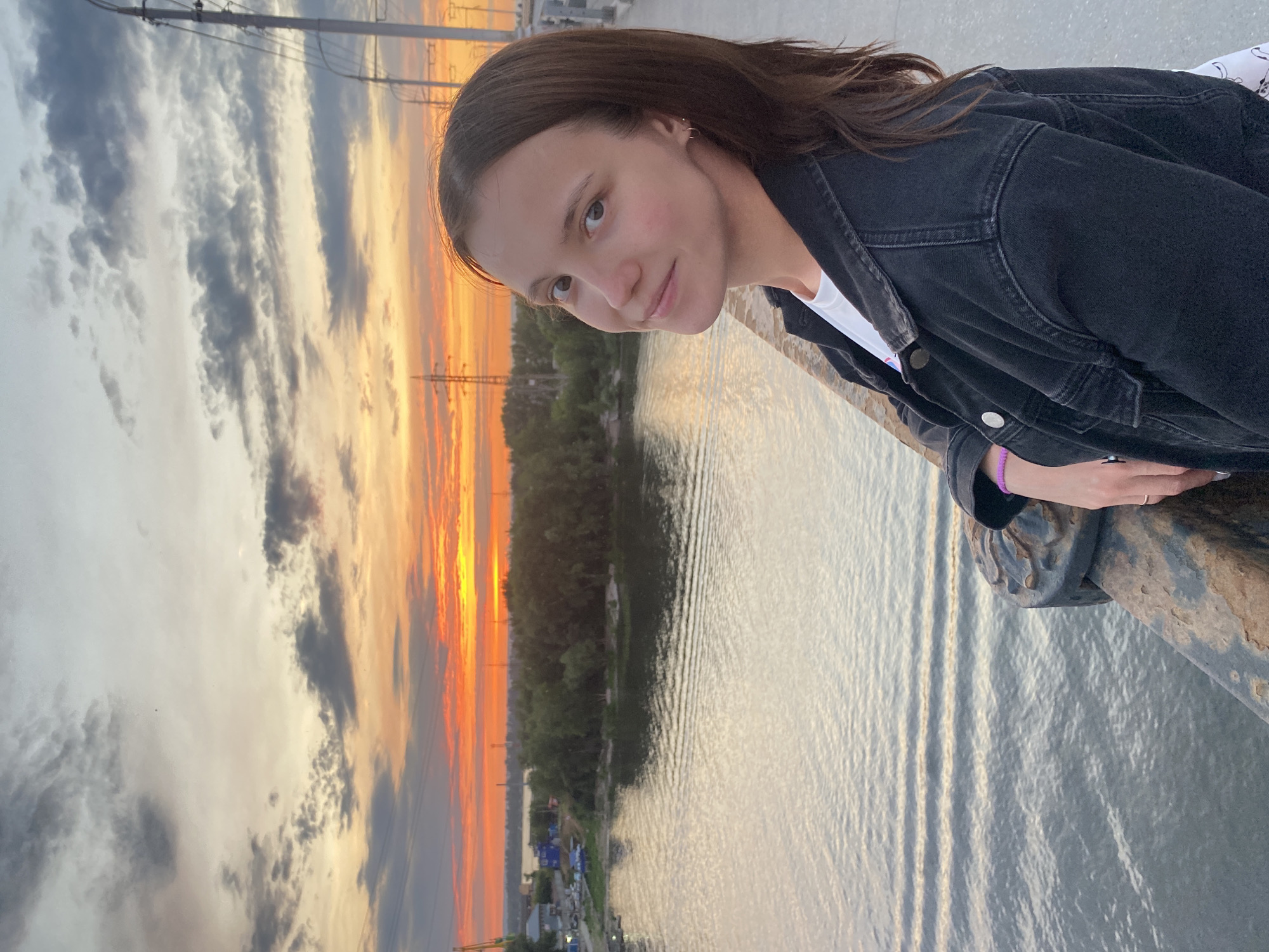 A young woman with brown hair and fair skin is photographed from the chest up, leaning on a rusty railing. She is wearing a white t-shirt and a black denim jacket. The woman is looking towards the camera with a slight smile. Behind her, a river flows with gentle ripples, and the opposite bank is lined with trees. The sky is filled with dramatic sunset clouds in shades of orange, yellow, and gray, with utility poles visible against the horizon. The scene appears to be on a bridge overlooking the Samara River in Novokuybyshevsk, Russia.