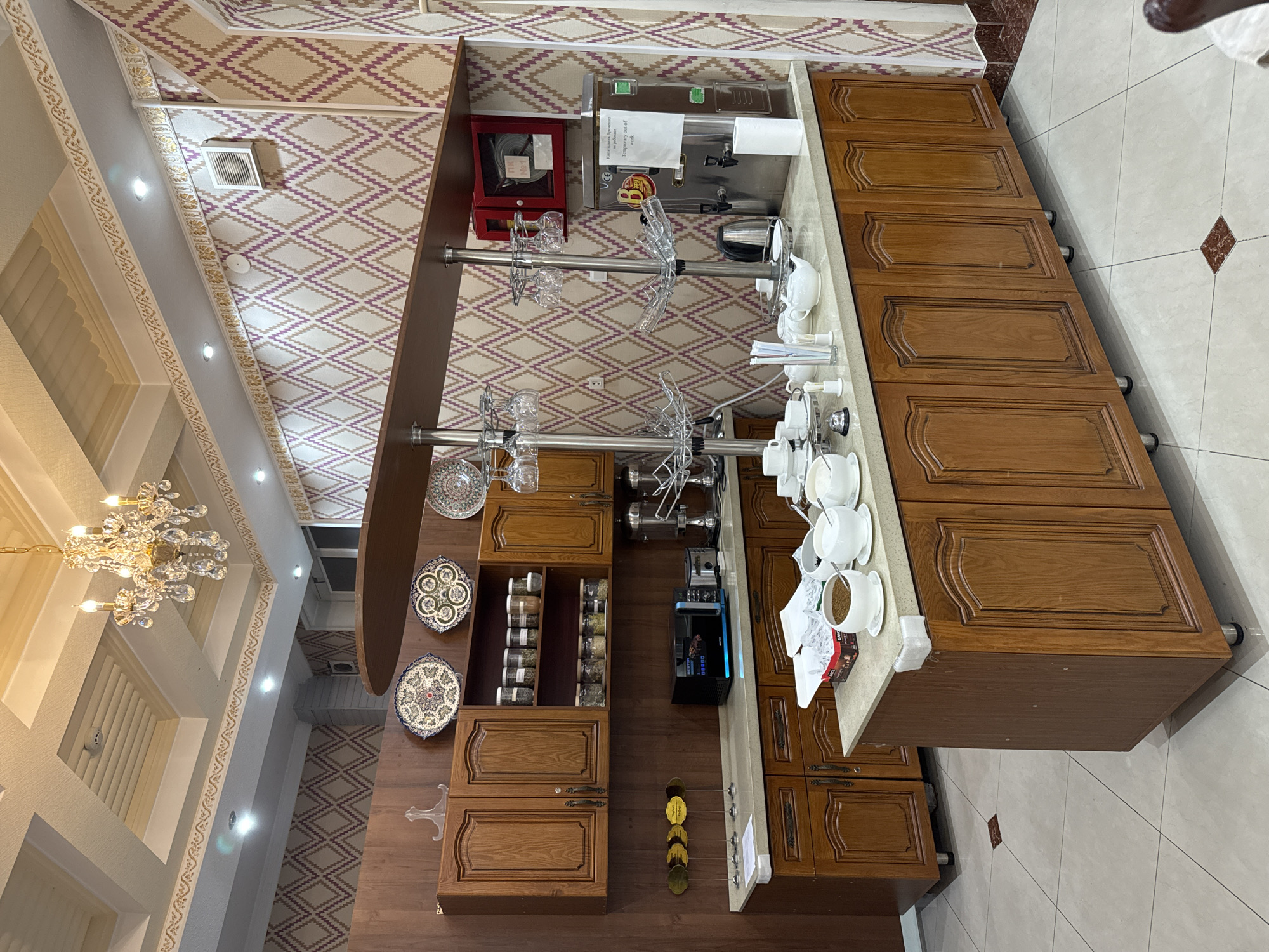The image shows a dining area in Samarkand City, Uzbekistan. It features a buffet-style setup with a tea and coffee station. There are wooden cabinets and countertops displaying various tea cups, teapots, and beverages. Plates are arranged decoratively on the wall. The ceiling is decorated with geometric patterns and a chandelier. The floor is tiled. There are no people visible in the scene.