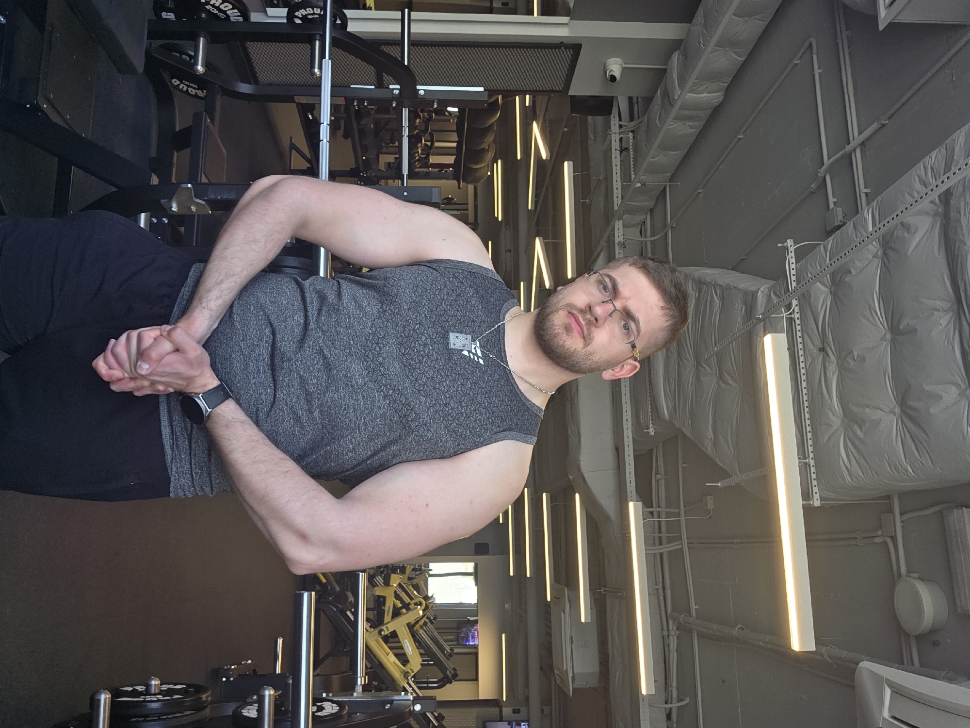 A man with short brown hair and glasses, wearing a gray tank top, black pants, and a watch, is in a gym. He has his hands clasped together. Various weightlifting machines are visible around him. The gym has a modern design with linear lighting fixtures. The location is possibly in Plovdiv, Bulgaria.